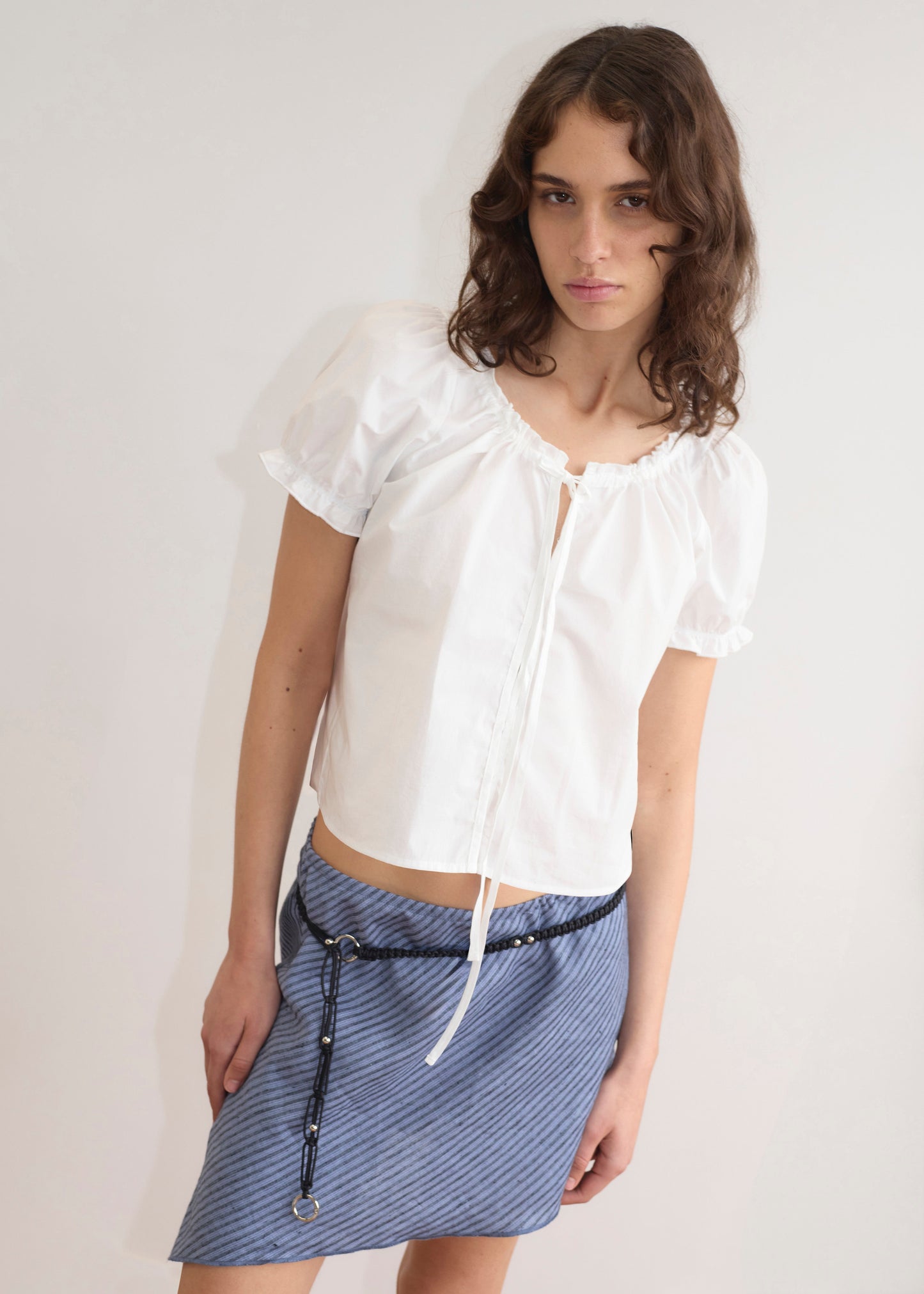 A woman with wavy brown hair wears the Close Shirt - White, featuring short sleeves and a gathered neckline, styled with a blue textured mini skirt and black belt, standing against a plain light-colored wall.