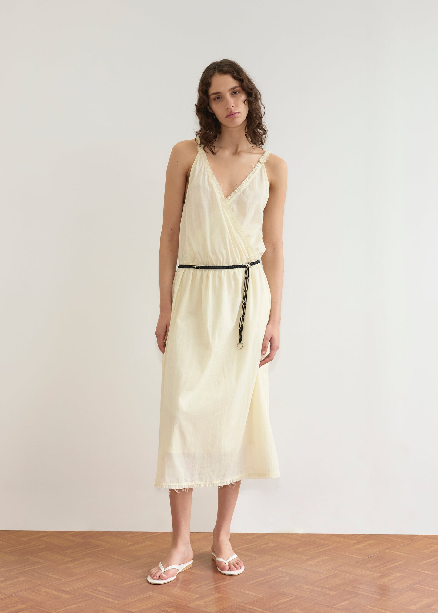 A woman with wavy brown hair stands indoors against a white wall, wearing the Crossed Dress - Soft Yellow Stripe by Deiji Studios with a black rope belt and white sandals. She has a neutral expression and slightly tilts her head.
