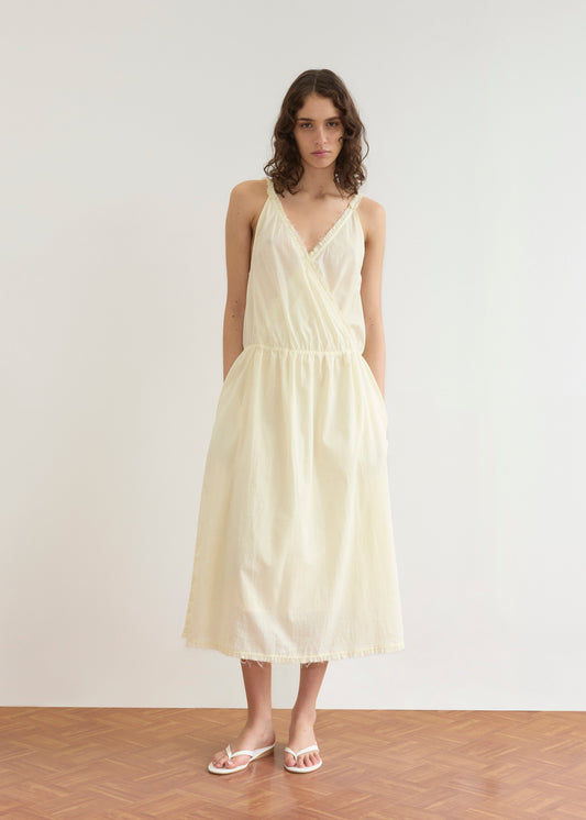 A woman with curly hair stands on a wooden floor against a plain white wall, wearing the Deiji Studios Crossed Dress in Soft Yellow Stripe and white sandals.