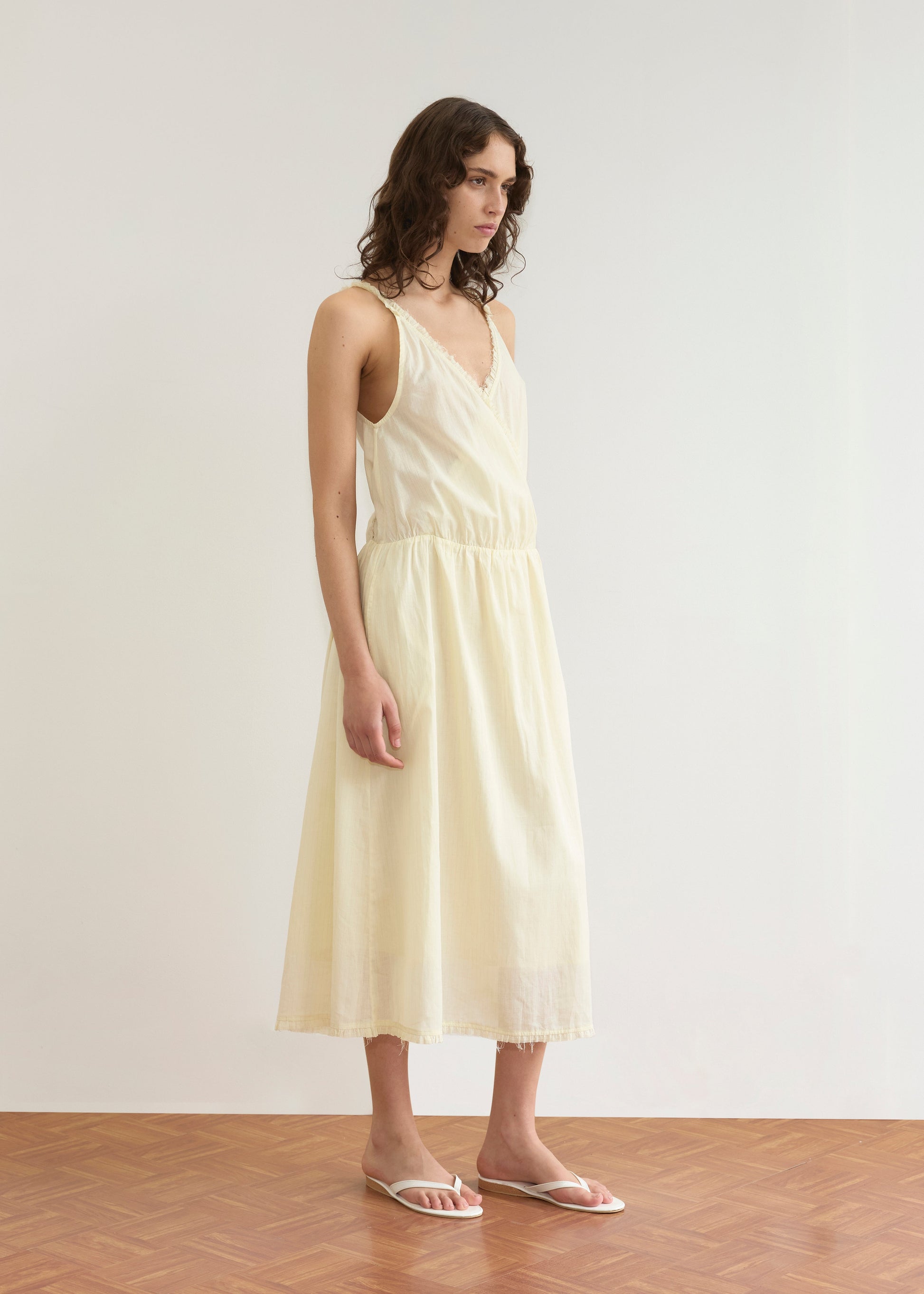 A woman with wavy brown hair stands indoors on a wooden floor, wearing the Crossed Dress - Soft Yellow Stripe from Deiji Studios, featuring a V-neck and thin straps, paired with white sandals. She looks slightly to the side.