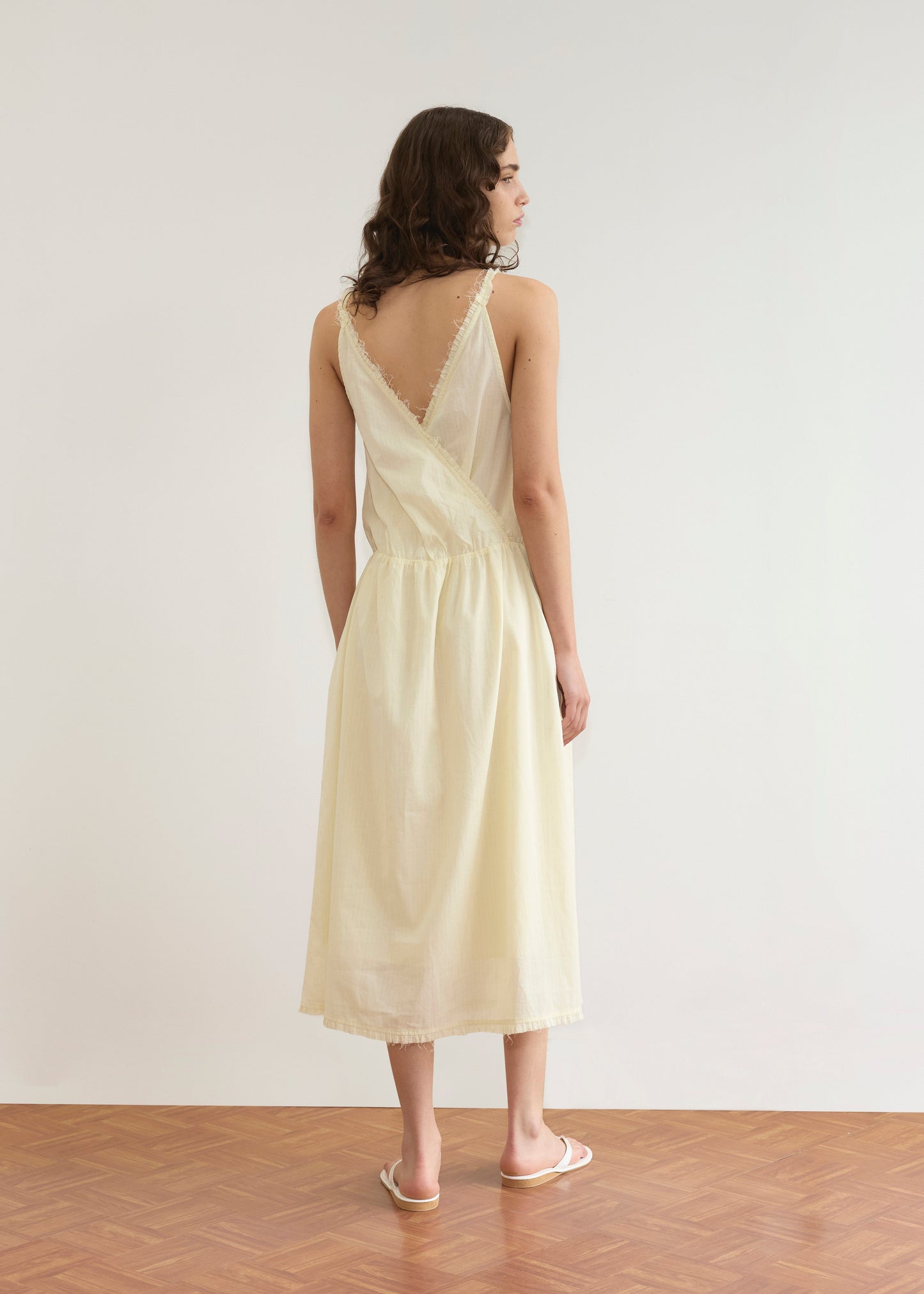 A woman with wavy brown hair and white sandals stands on a wooden floor, facing away, wearing the Crossed Dress - Soft Yellow Stripe by Deiji Studios. It features a sleeveless design, deep V-back, frayed edges, and a plain white background.