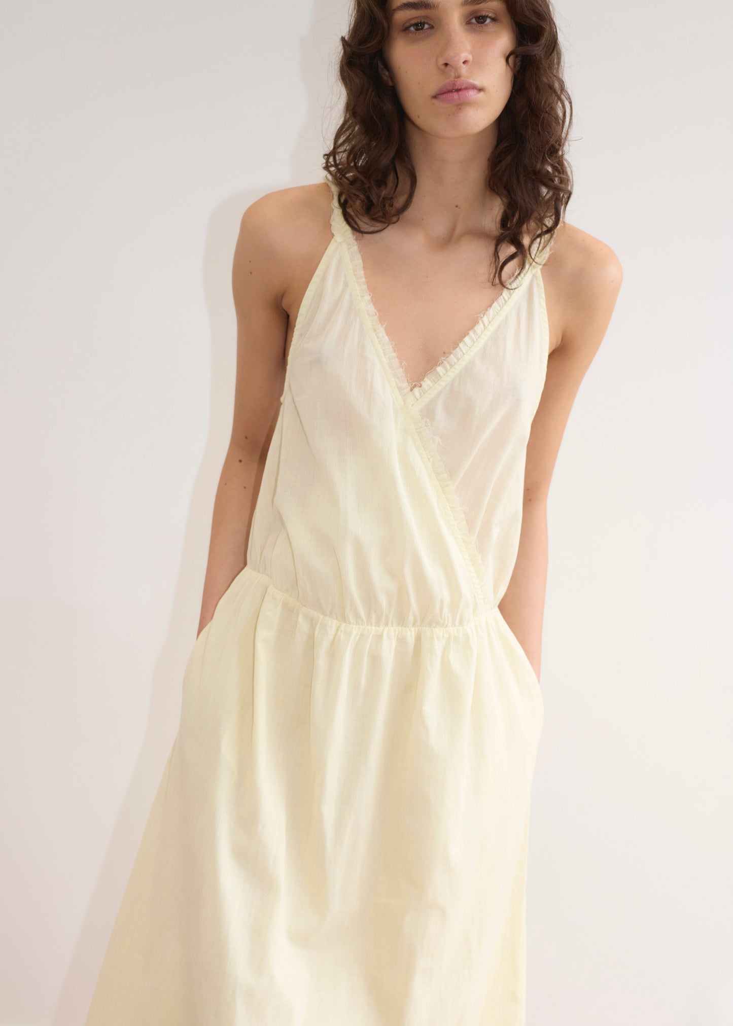 A model wears the Crossed Dress - Soft Yellow Stripe from Deiji Studios, a cream sleeveless midi with deep V-neckline, ruffle trim, elasticated waist, full skirt, side pockets, and light airy fabric. Minimalist white background.