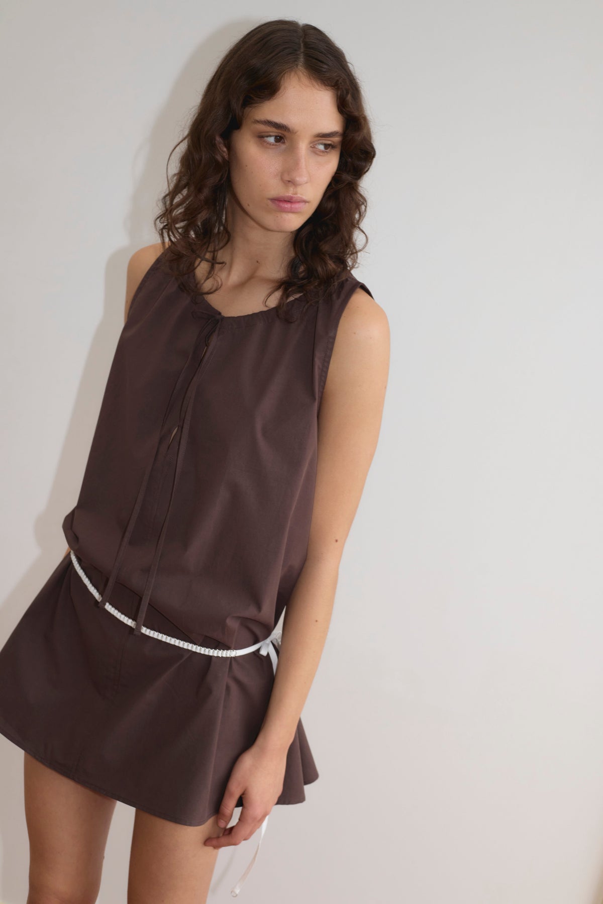 A person with wavy brown hair wears the sleeveless Drawcord Dress - Sepia by Deiji Studios, featuring an a-line silhouette and white belt, standing against a plain light wall and looking slightly left with a neutral expression.