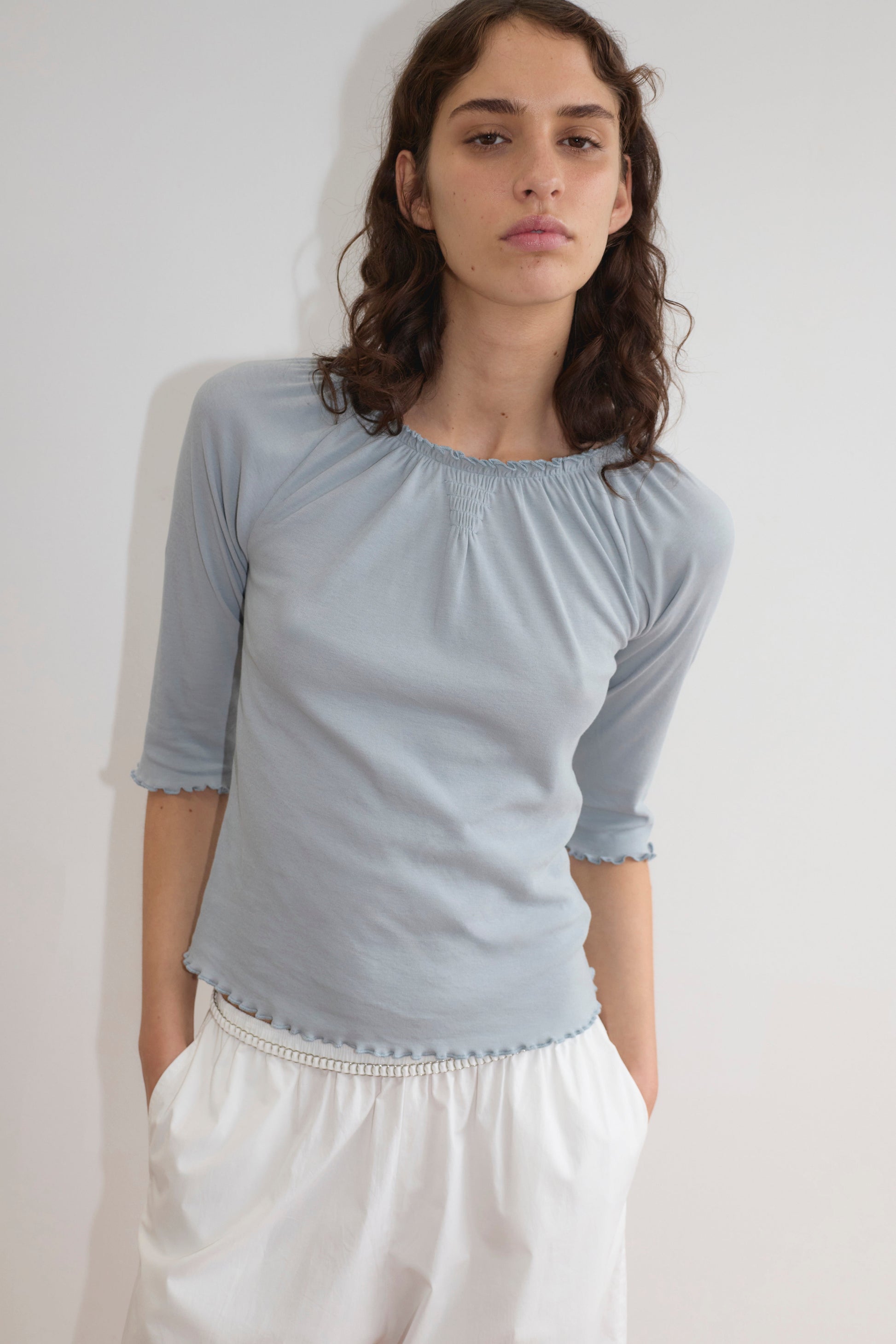 A person with wavy brown hair wears the Deiji Studios Cotton Flounce 3/4 Sleeve top in baby blue and white pants, standing against a plain white background with hands in pockets, looking at the camera with a neutral expression.