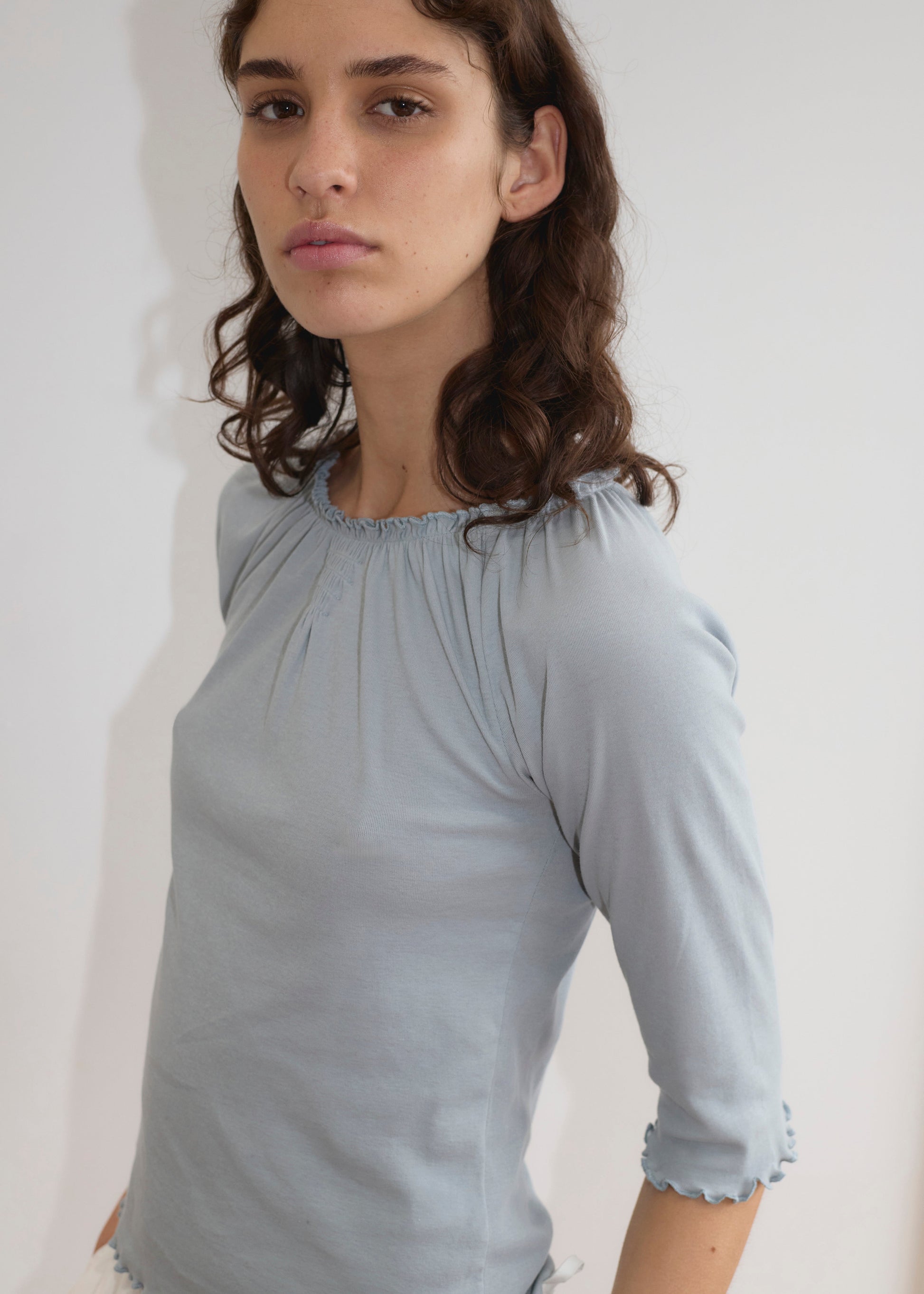 A person with wavy brown hair wears the Cotton Flounce 3/4 Sleeve in Baby Blue, standing against a plain white background and looking at the camera with a neutral expression.