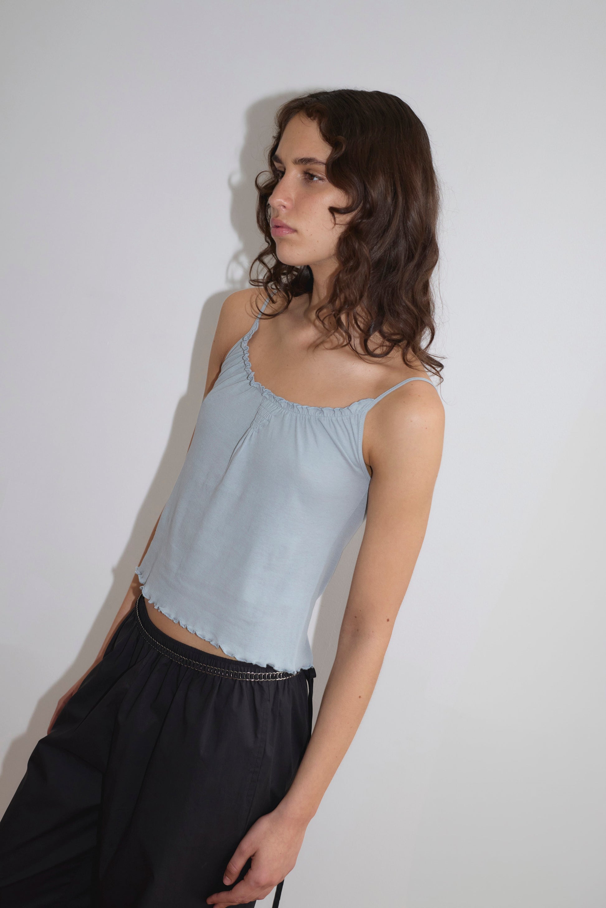A young person with wavy brown hair wears a Cotton Flounce Tank in baby blue and black pants, standing against a plain white wall and looking slightly left with a neutral expression.