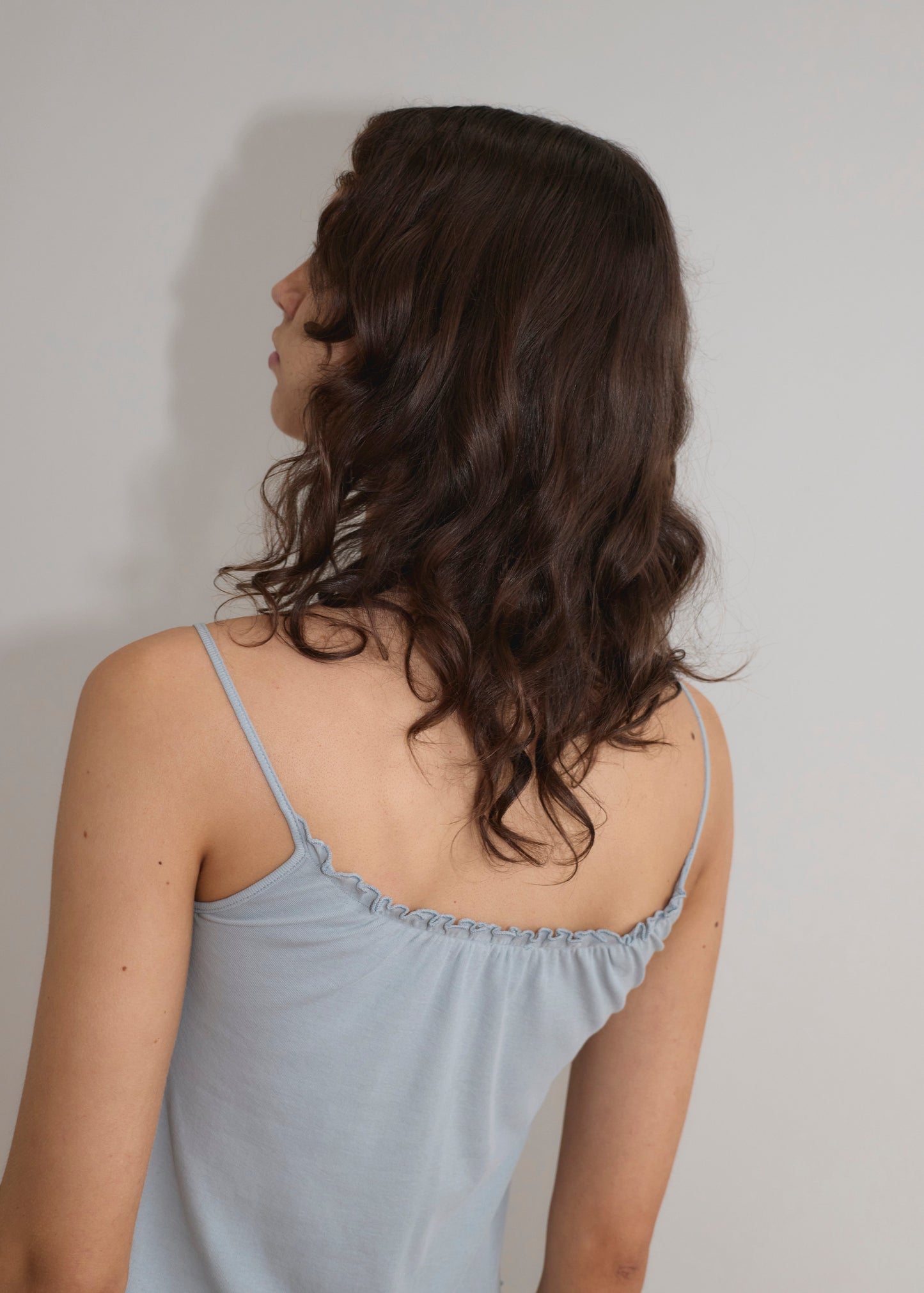 A person with wavy brown hair stands with their back to the camera, wearing a Cotton Flounce Tank - Baby Blue against a plain light background.