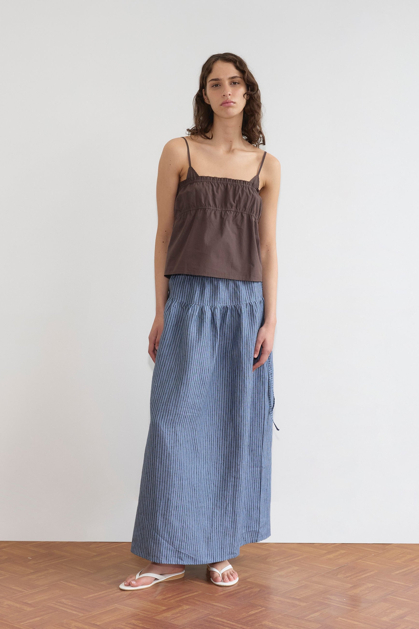 A woman with long curly hair stands against a white wall in the Deiji Studios Tri Cami - Sepia top, a relaxed blue and white striped maxi skirt, and white sandals. She appears neutral-faced on a wooden floor.