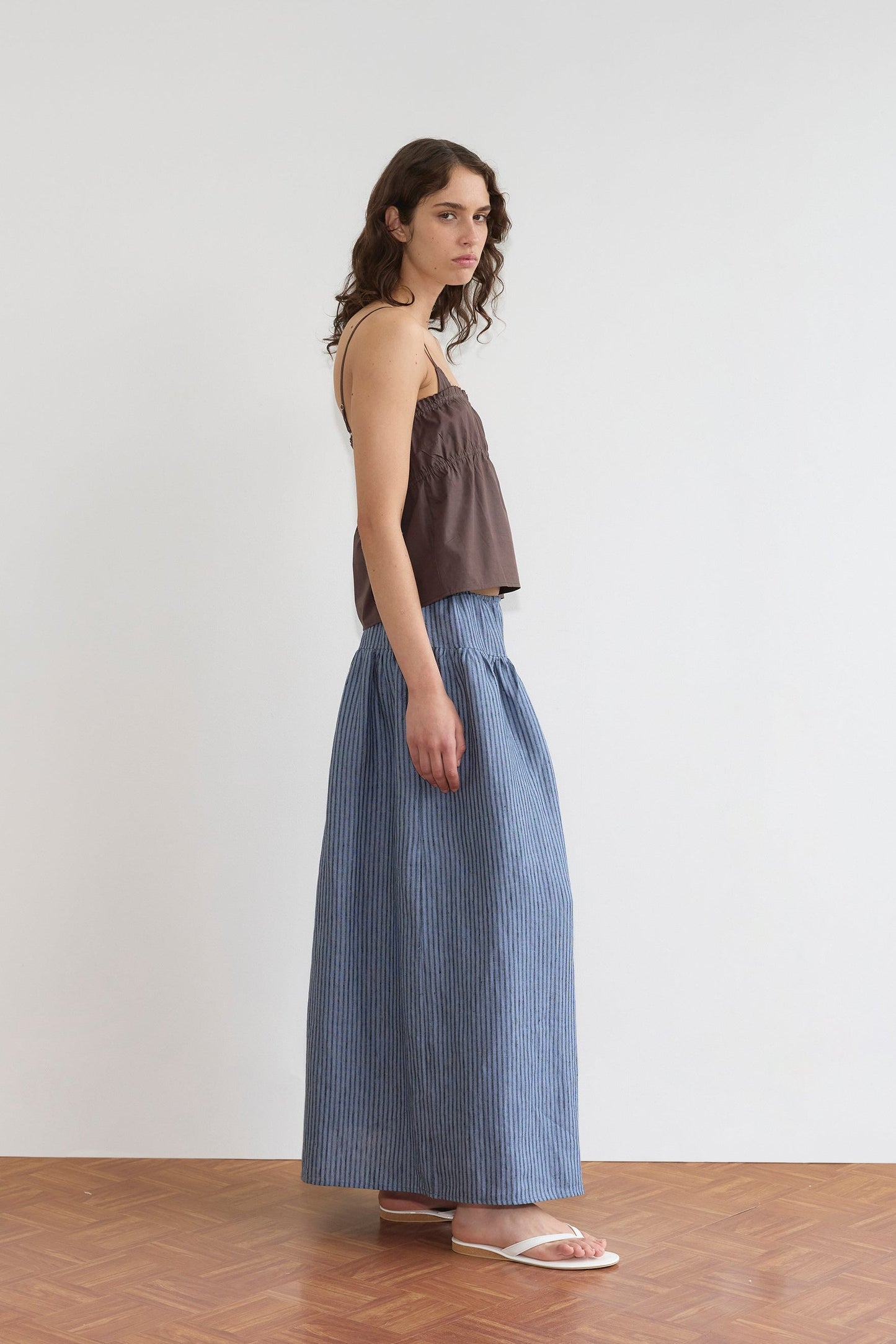 A person with long, wavy hair wears a relaxed silhouette—a brown Tri Cami - Sepia from Deiji Studios paired with a blue and white striped maxi skirt and white sandals, standing sideways on a wooden floor against a plain white wall.