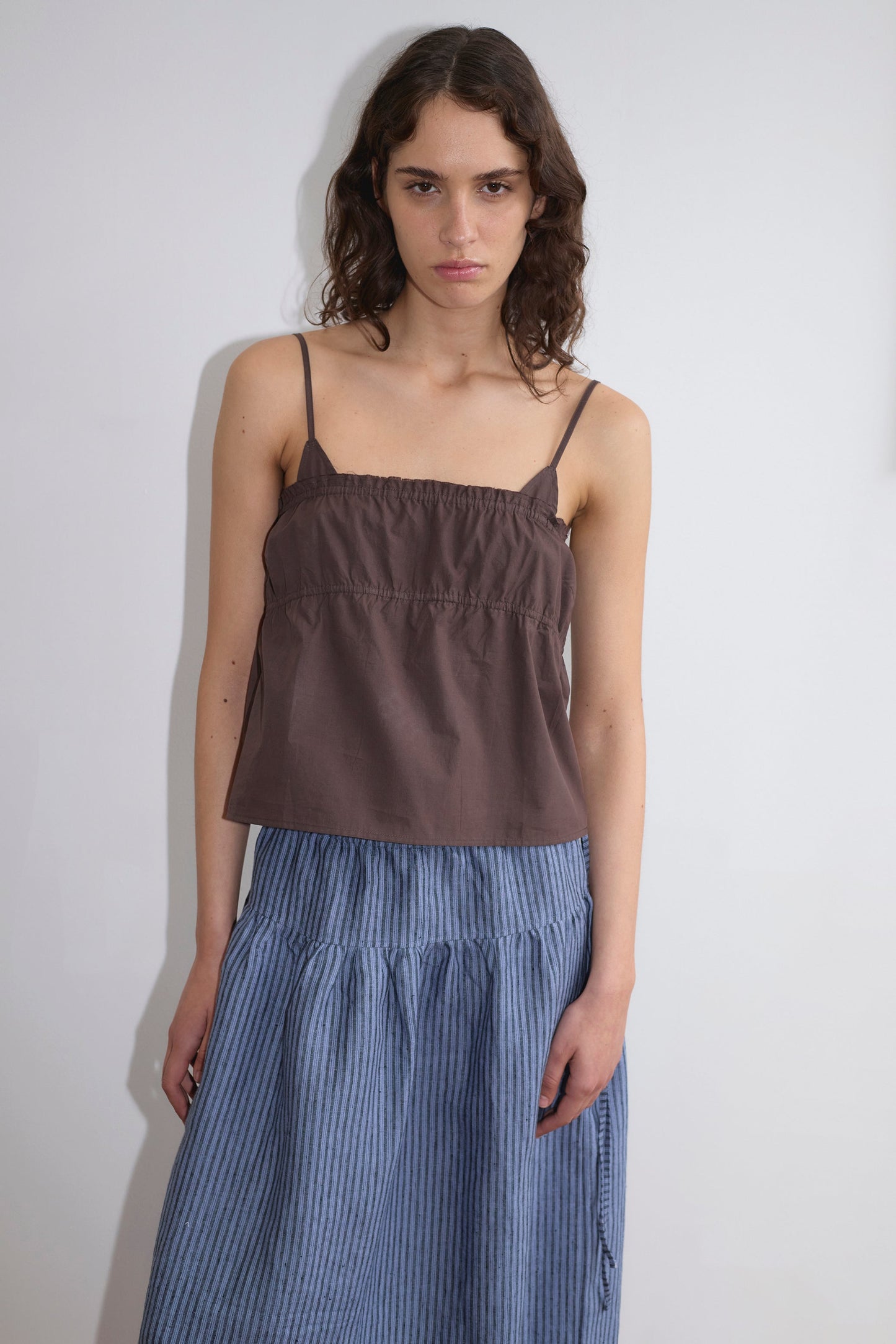 A person with wavy brown hair stands against a white wall, wearing the Tri Cami - Sepia by Deiji Studios and a blue and white striped skirt, showcasing a relaxed silhouette while gazing neutrally at the camera.