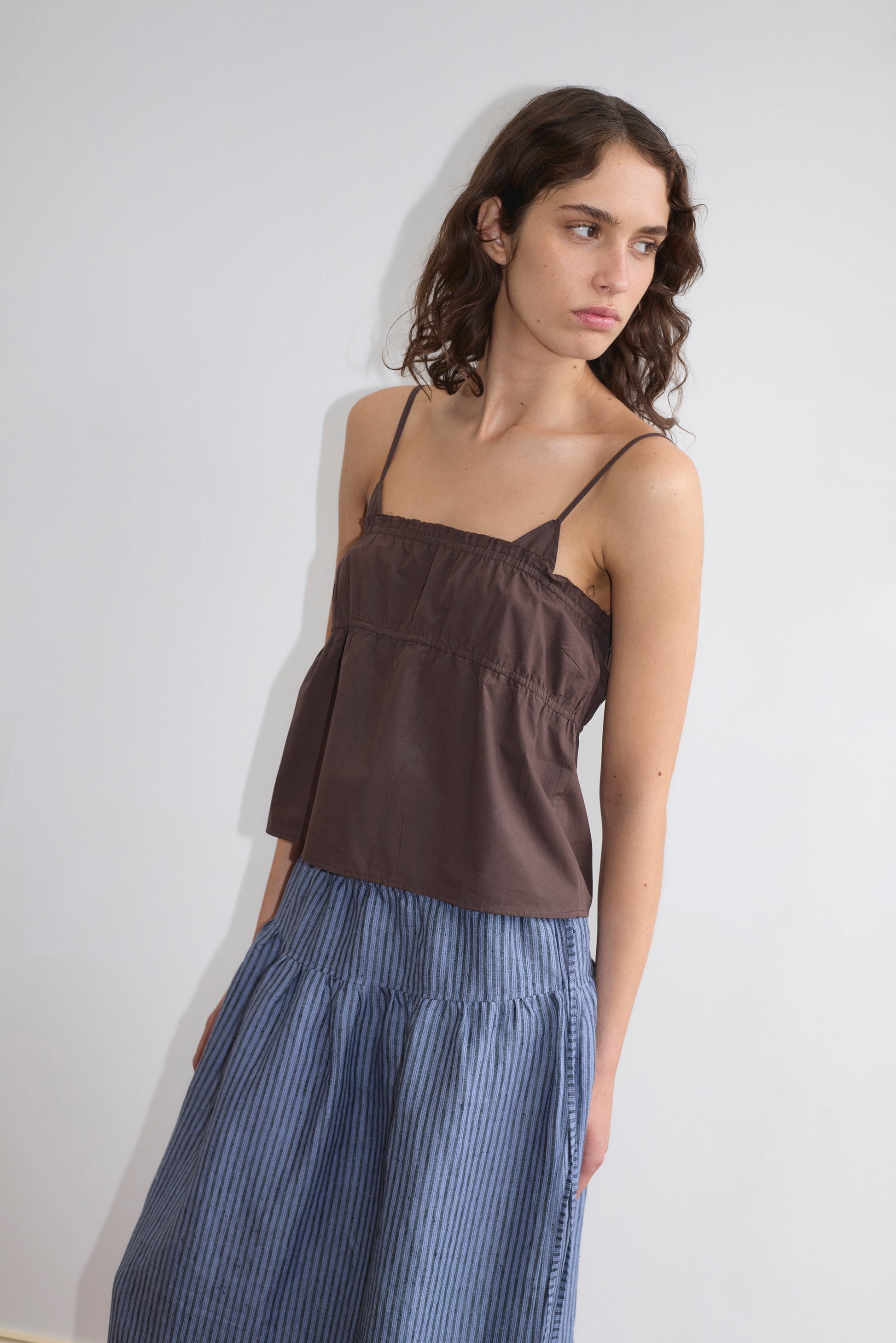A woman with wavy brown hair wears the Tri Cami in Sepia by Deiji Studios and a blue, high-waisted pleated skirt. She stands against a plain white wall, looking to her left in a relaxed pose.