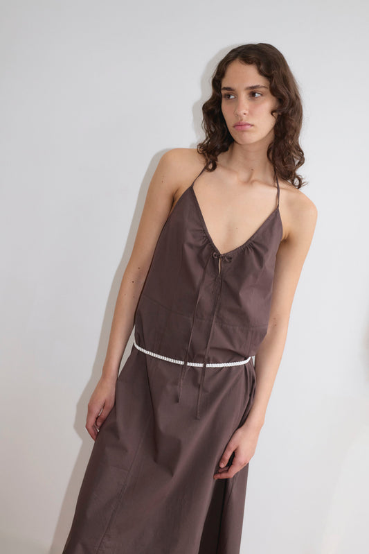 A woman with wavy brown hair wears the Seamed Halter Dress, a sleeveless deep V-neck brown dress with a thin white belt, standing against a plain white background.