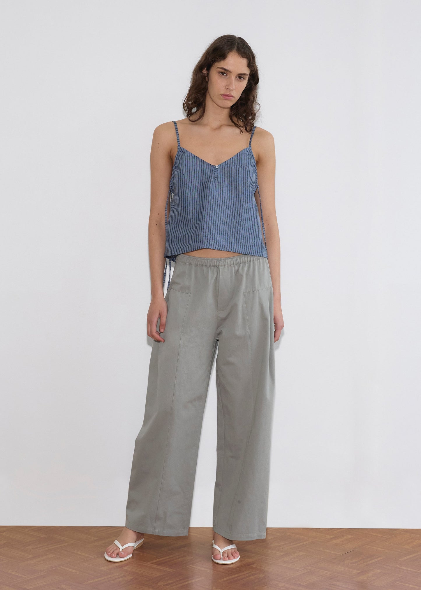 A woman stands indoors wearing the V Neck Cami - Azure Stripe from Deiji Studios, paired with loose gray pants and white sandals on a wooden floor against a plain white background.