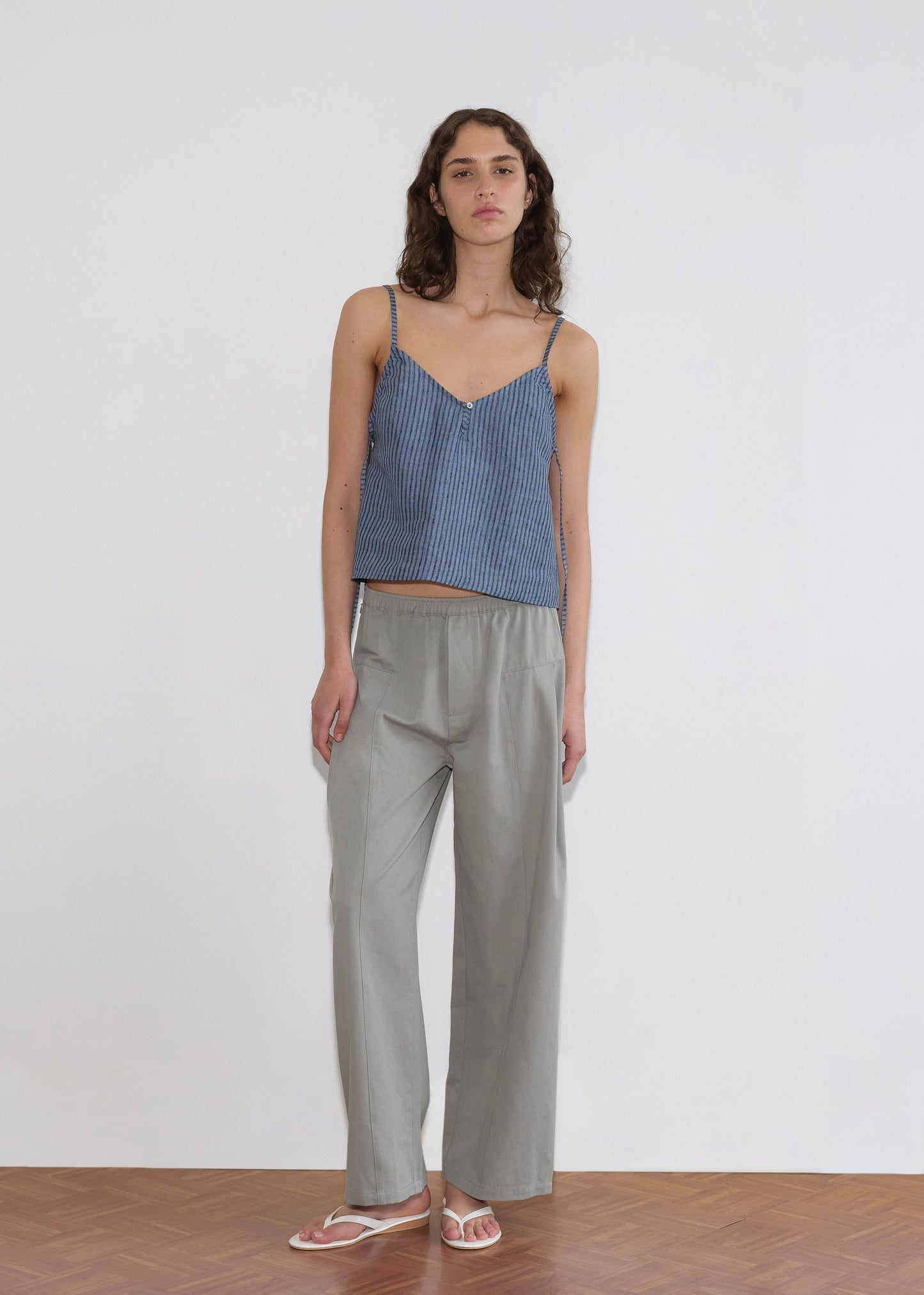 A woman with curly hair and a neutral expression stands indoors against a white wall, wearing the Deiji Studios V Neck Cami - Azure Stripe, loose light gray pants, and white flip-flops.