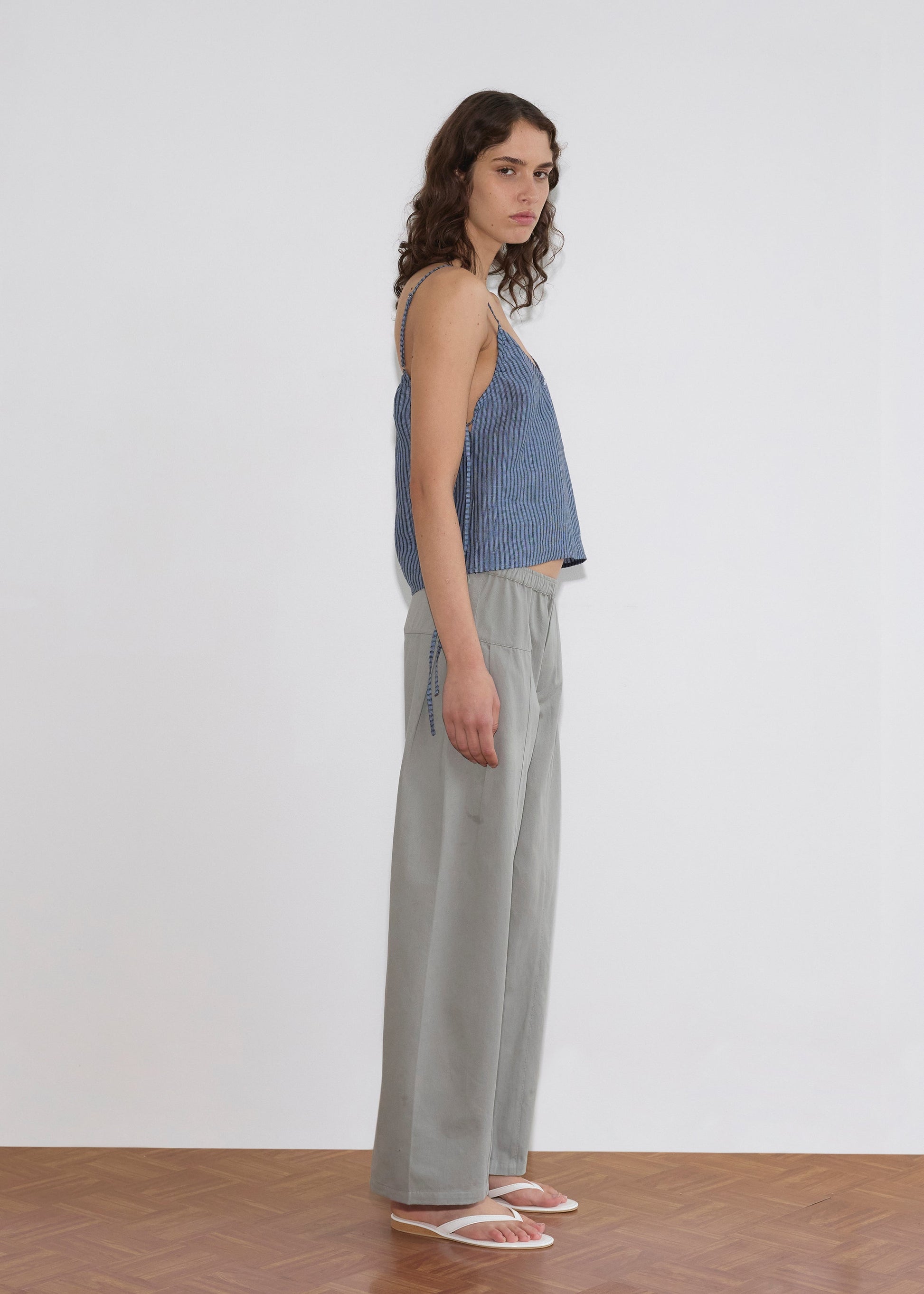 A person with wavy brown hair stands sideways in a Deiji Studios V Neck Cami - Azure Stripe, paired with loose light gray pants and white sandals, set against a plain white wall and wooden floor.