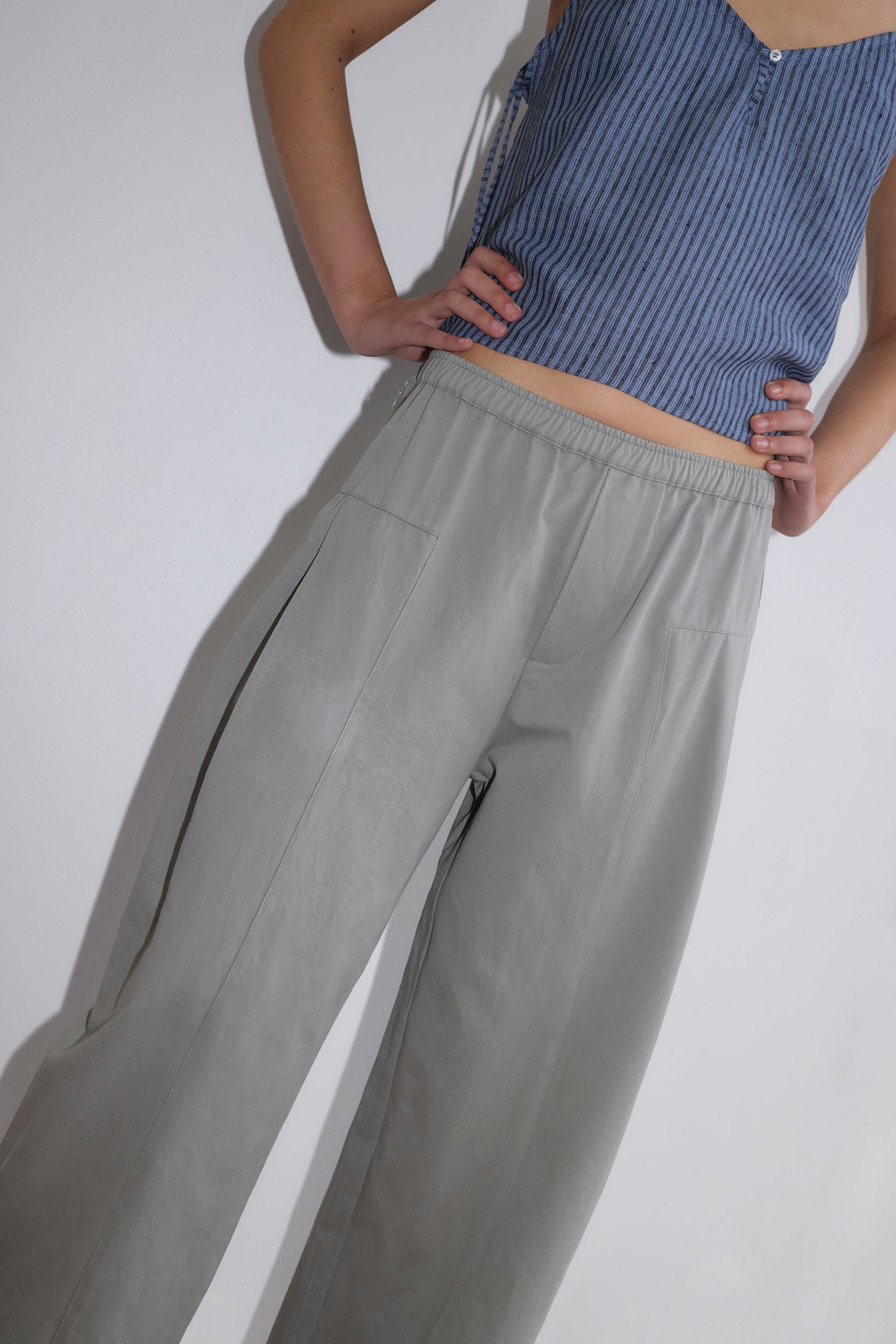A person stands with hands on hips, wearing the V Neck Cami - Azure Stripe by Deiji Studios and loose, light gray pants. Their face is not visible.