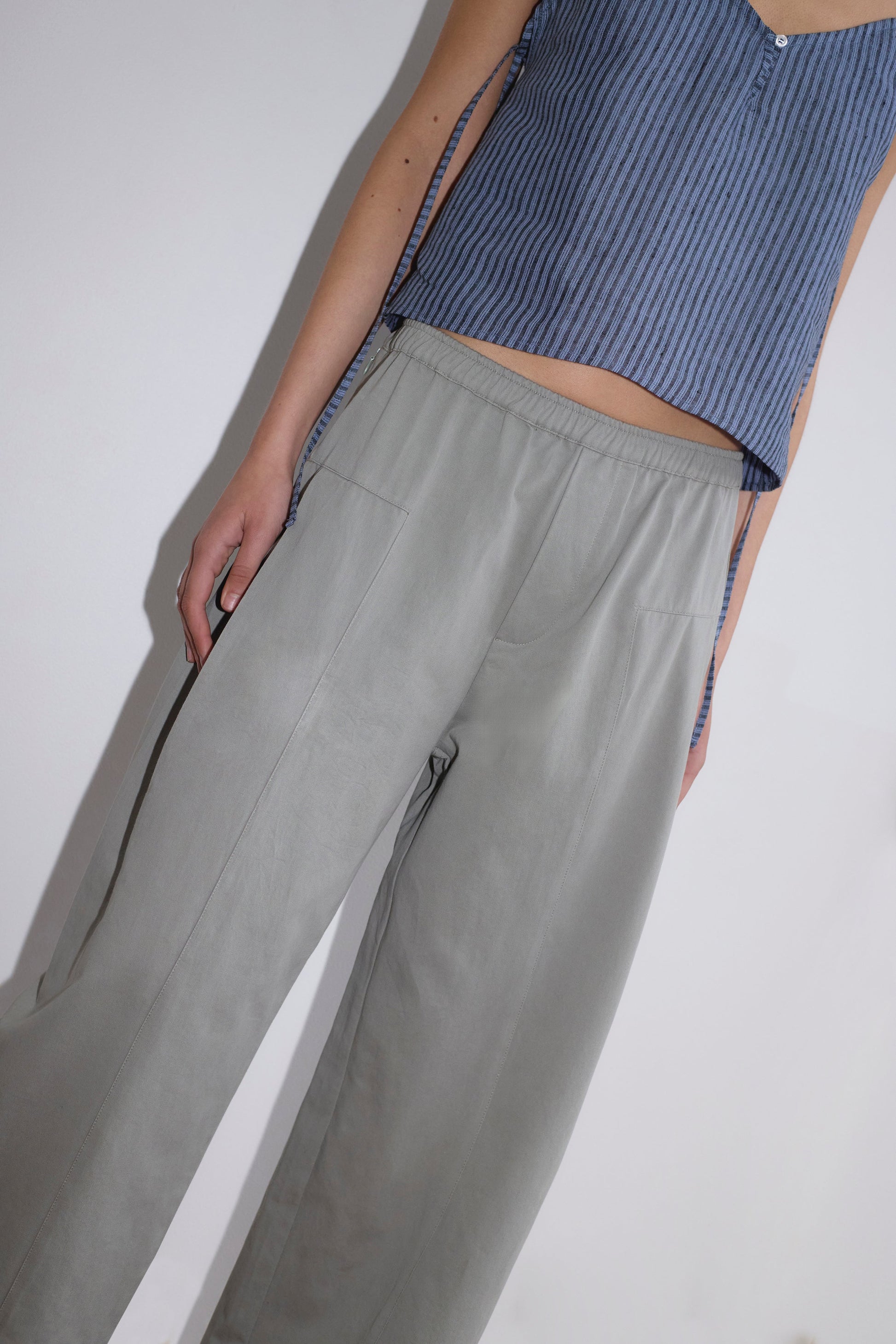A person wearing the Pleat Pant - Sage Grey by Deiji Studios and a sleeveless blue pinstriped top stands against a plain white background, showing only their lower torso, one arm, and legs.