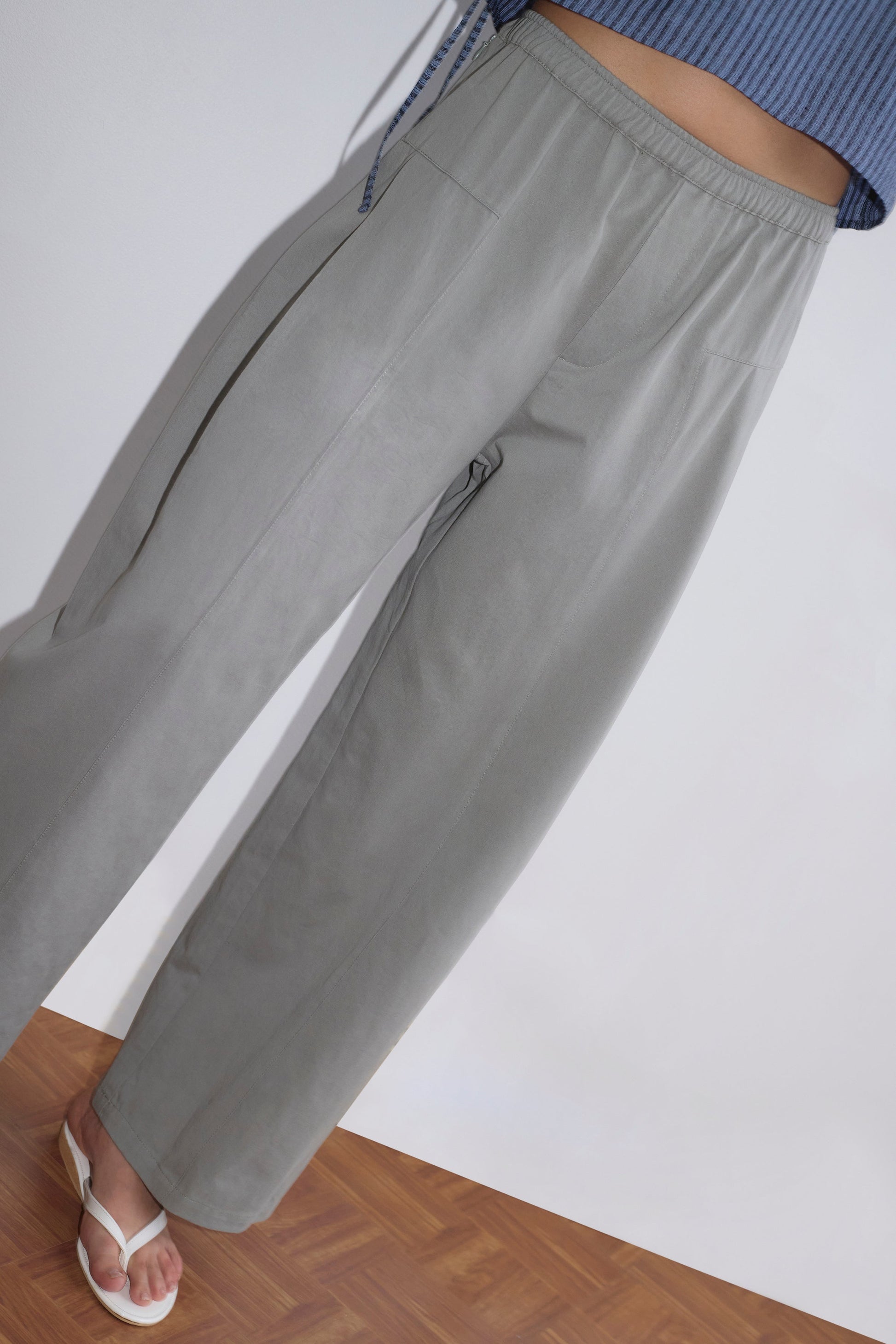 A person stands on a wooden floor against a plain white wall, wearing Deiji Studios' Pleat Pant in Sage Grey, a blue ribbed top, and white heeled sandals. The person's face is not visible.