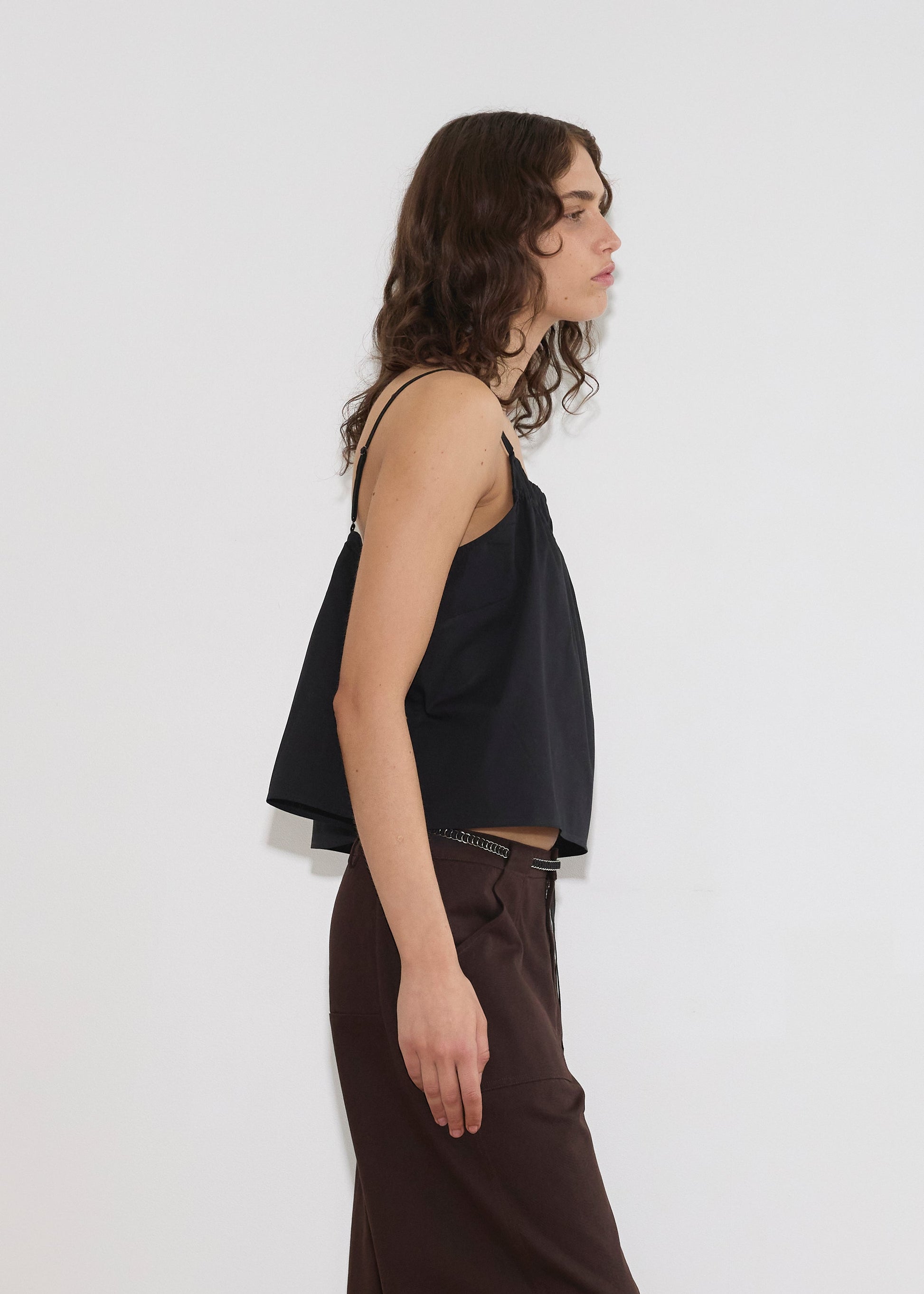A person with wavy brown hair stands in profile against a white background, wearing the Double Layer Top - Black by Deiji Studios and dark brown pants.
