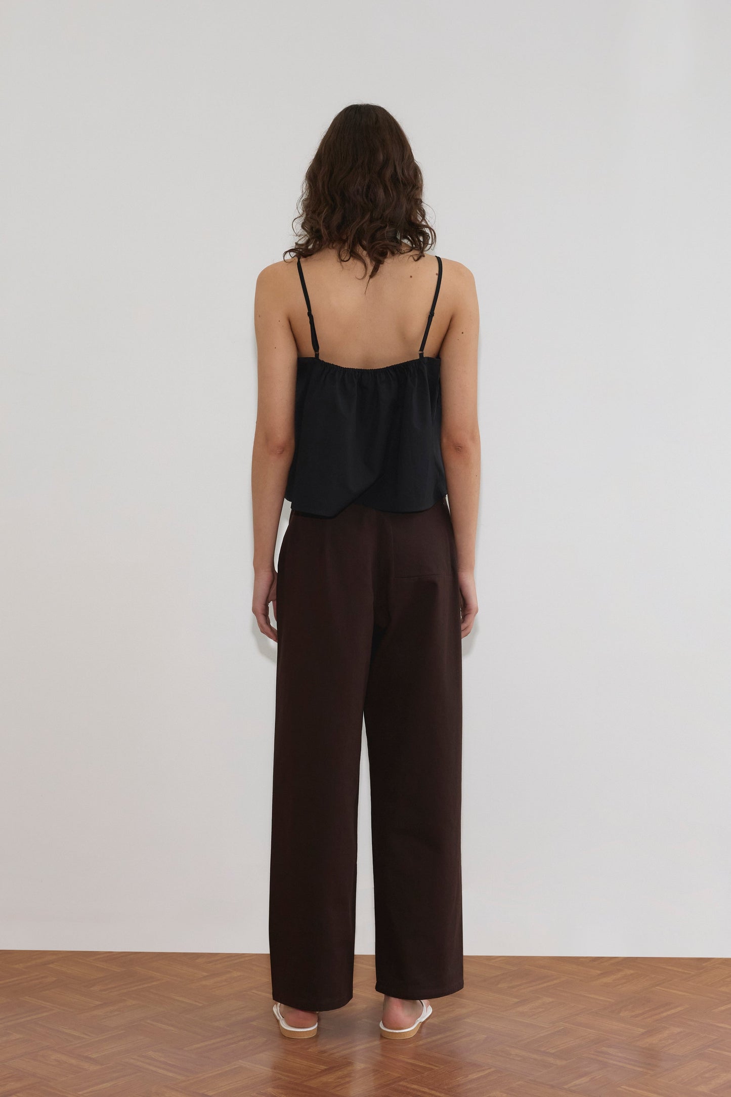 A person with wavy brown hair stands facing a white wall, wearing a black sleeveless top, Pocket Jean - Dark Brown by Deiji Studios, and white heeled sandals on a wooden floor.