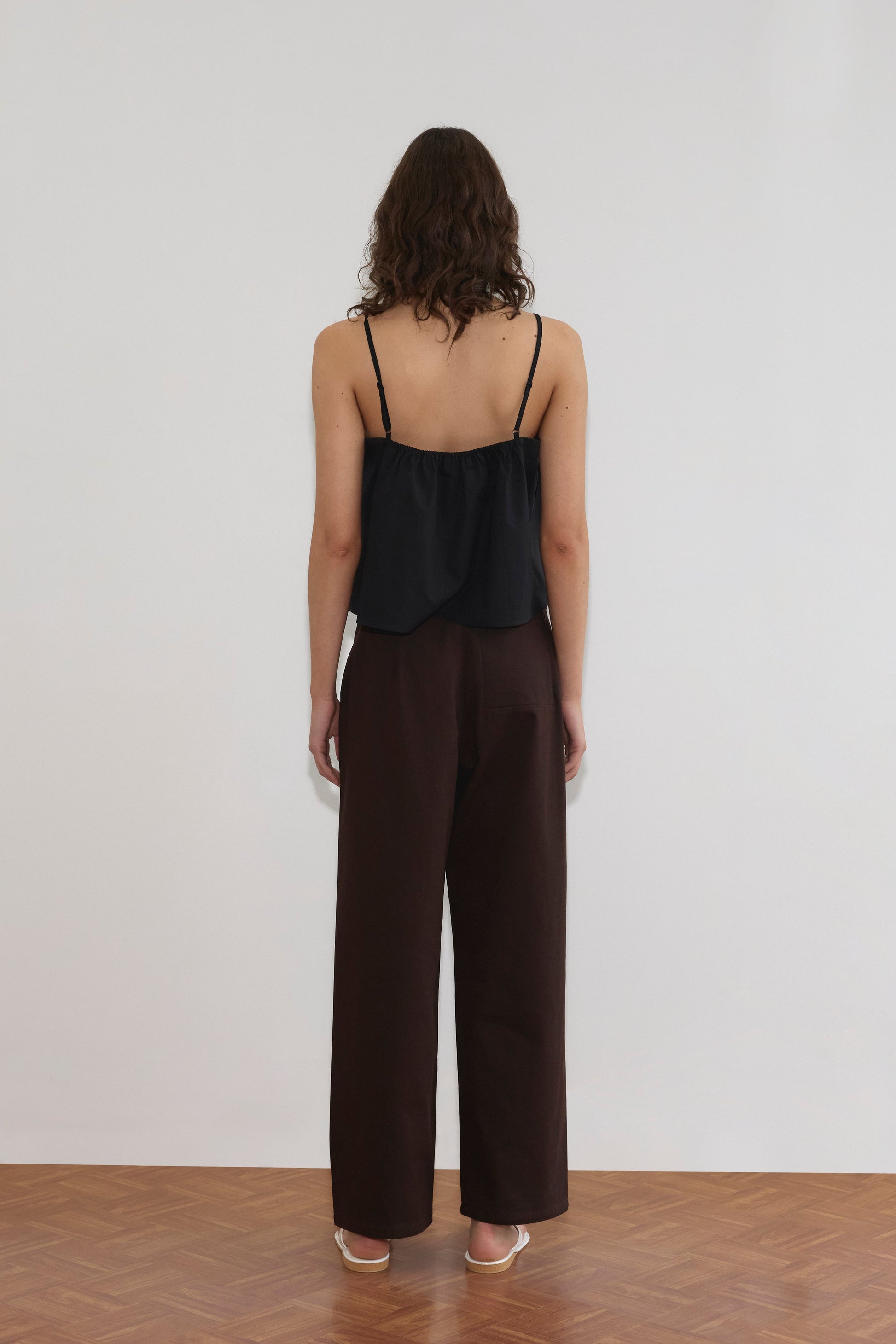 A person with wavy brown hair stands facing a white wall, wearing a black sleeveless top, Pocket Jean - Dark Brown by Deiji Studios, and white heeled sandals on a wooden floor.