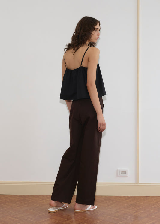A person with wavy brown hair stands sideways, wearing the Double Layer Top - Black by Deiji Studios, wide-legged dark brown pants, and white sandals in a minimalist room with wooden floors and white walls.