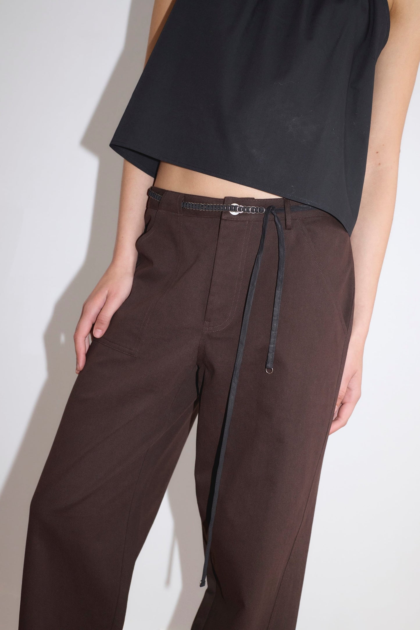 A person wearing a loose black sleeveless top and the Pocket Jean - Dark Brown by Deiji Studios with a thin black belt, standing against a plain light-colored background.