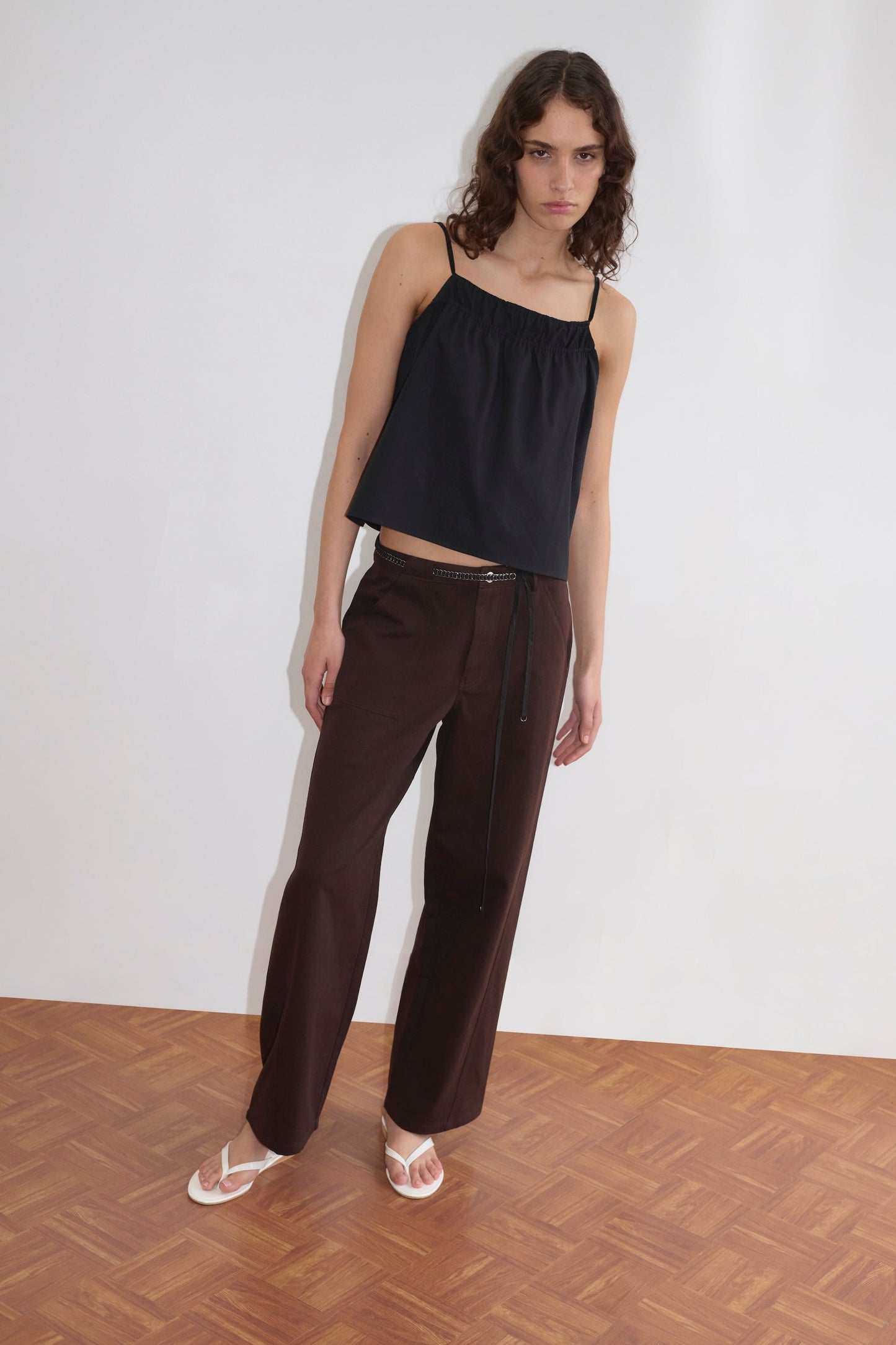 A person with wavy brown hair wears a loose black spaghetti strap top, Pocket Jean - Dark Brown by Deiji Studios with a belt, and white sandals, standing on a wooden parquet floor against a plain white wall.