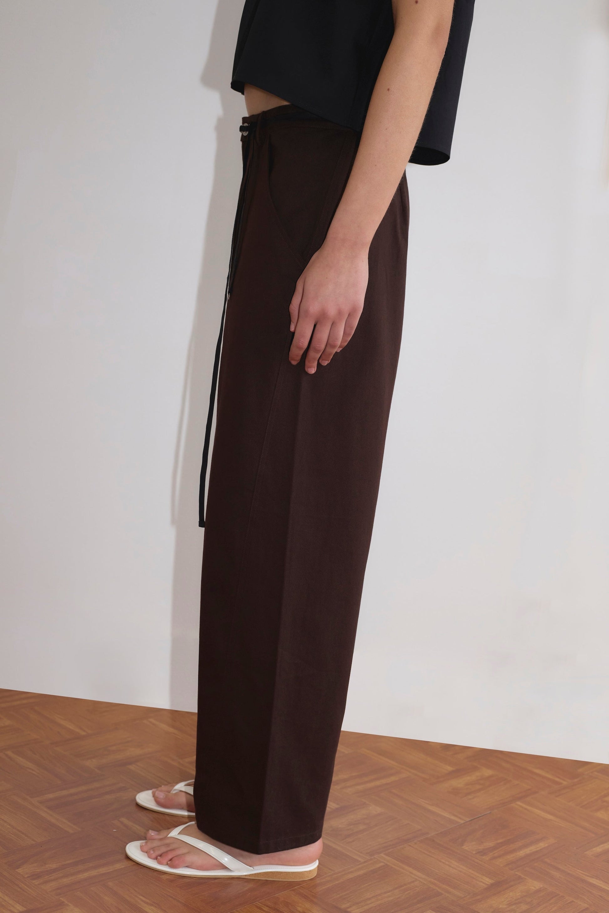 A person stands sideways on a wooden floor, wearing the Pocket Jean - Dark Brown from Deiji Studios with a black top and white sandals. The background is a plain light-colored wall.