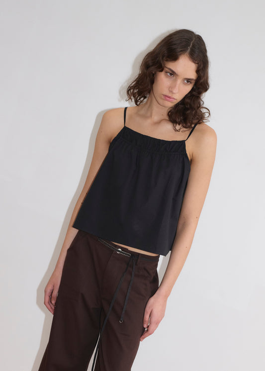 A woman with wavy brown hair wears the Double Layer Top - Black by Deiji Studios and dark brown pants, standing against a plain white wall and looking slightly downward with a neutral expression.