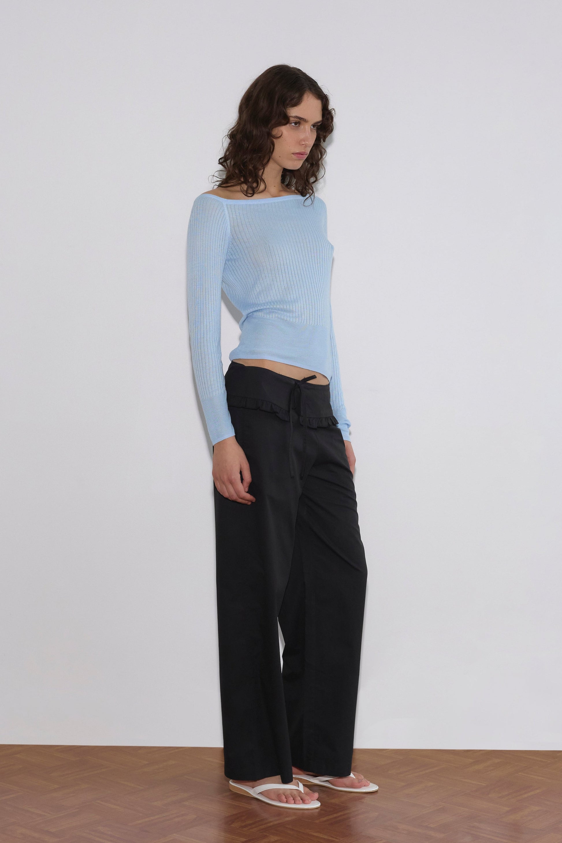 A person with wavy brown hair stands against a white wall on a wooden floor, wearing a light blue off-shoulder ribbed top, Flounce Belt Pant in black, and white flip-flops.