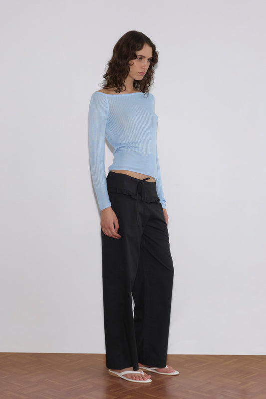A person with wavy brown hair stands against a white wall on a wooden floor, wearing a light blue off-shoulder ribbed top, Flounce Belt Pant in black, and white flip-flops.