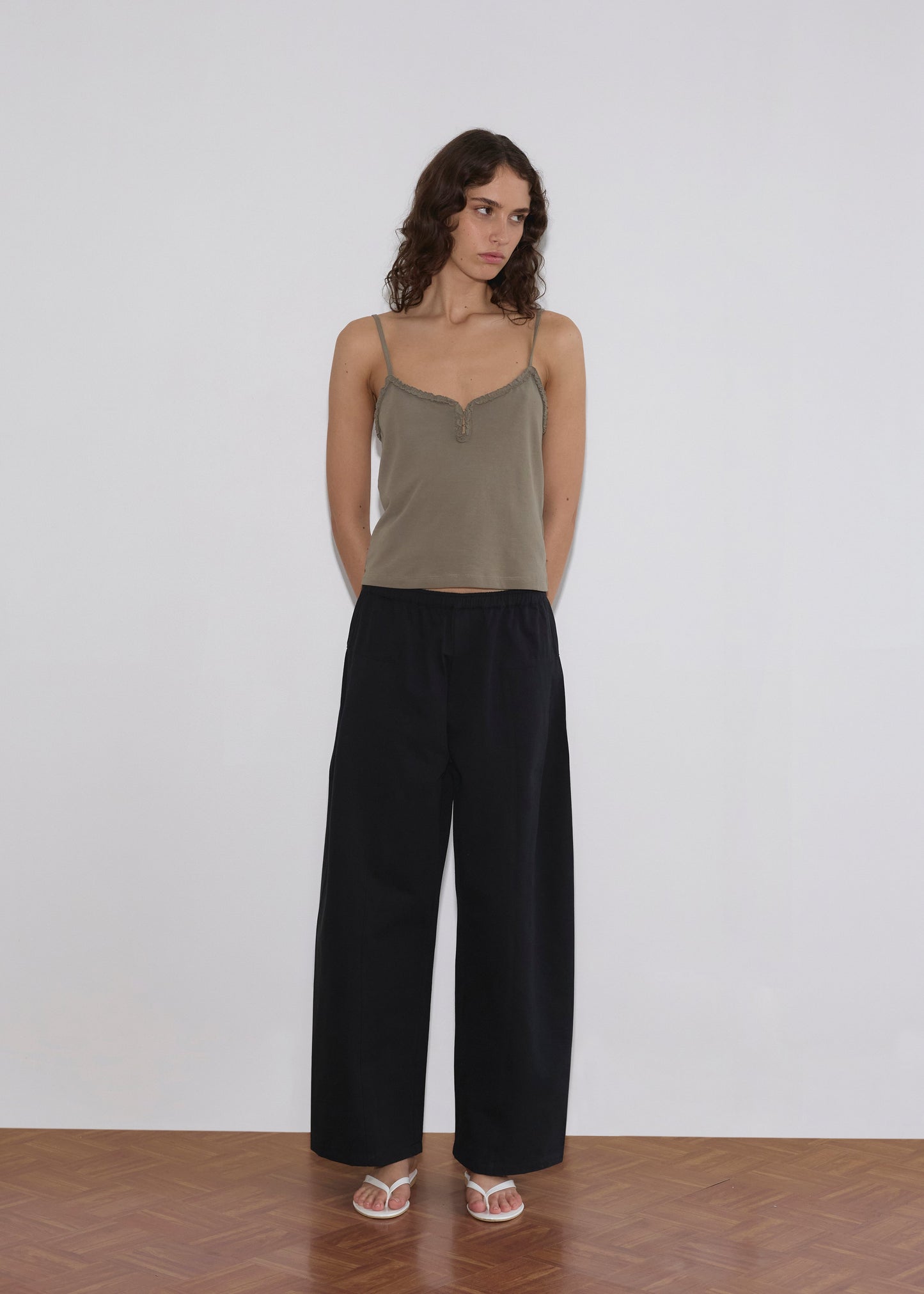 A woman with wavy brown hair stands against a plain white wall, wearing a khaki camisole, Deiji Studios Pleat Pant in Black, and white sandals. Her hands are behind her back as she gazes slightly to the side.