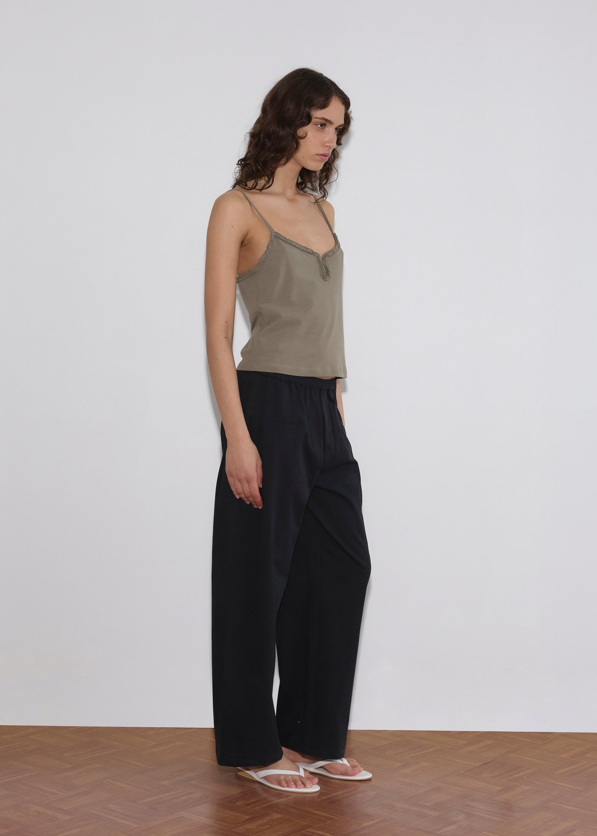 A person stands sideways against a white wall, wearing the Clasp Singlet - Khaki by Deiji Studios with adjustable straps, loose black pants, and transparent sandals on a wooden floor.