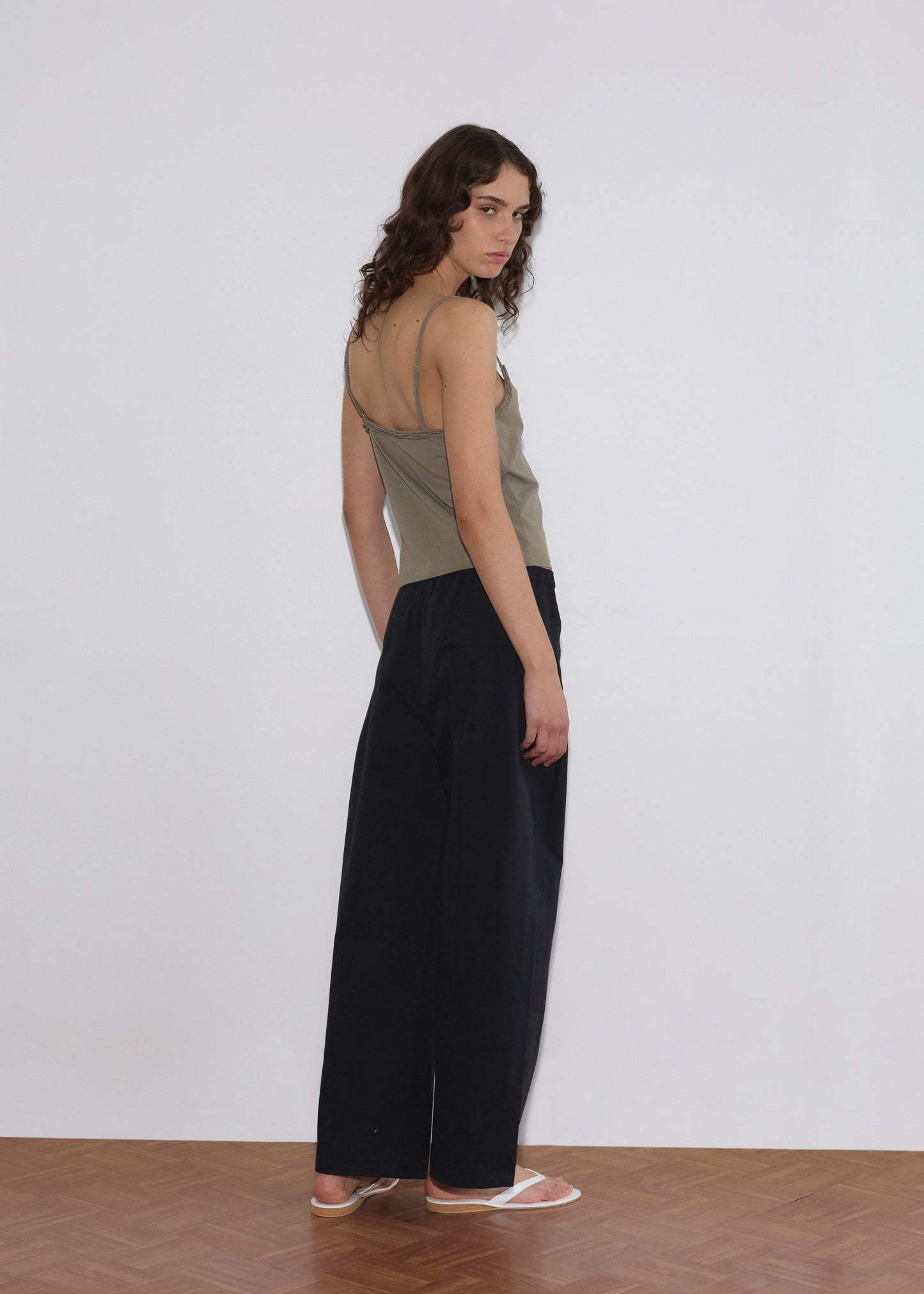 A person with long curly hair stands indoors against a white wall, wearing a taupe tank top, Pleat Pant - Black, and white sandals, looking over their shoulder toward the camera.