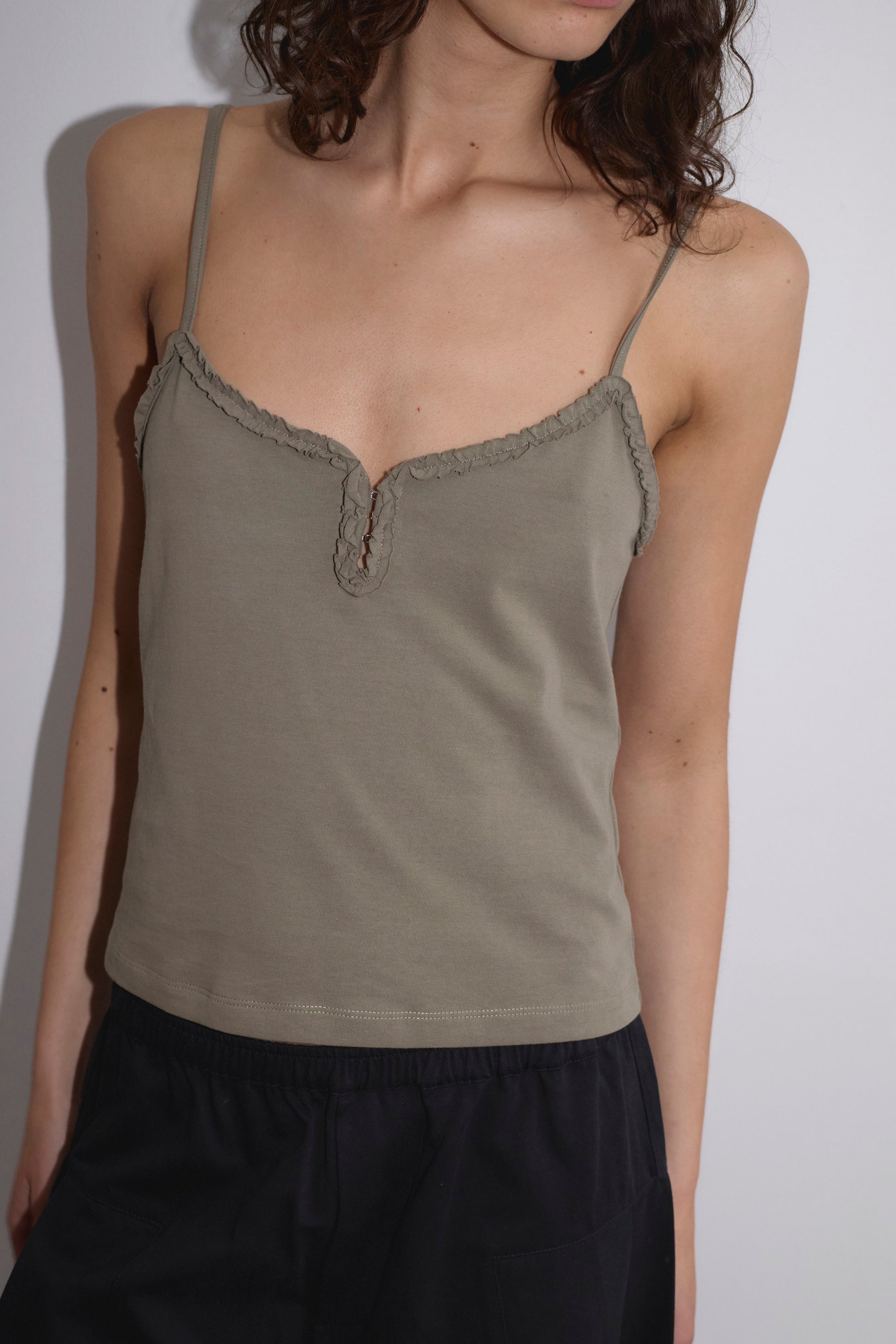A person with wavy brown hair wears the light olive green Clasp Singlet featuring thin straps and a scalloped neckline, paired with black pants, standing against a plain white wall.