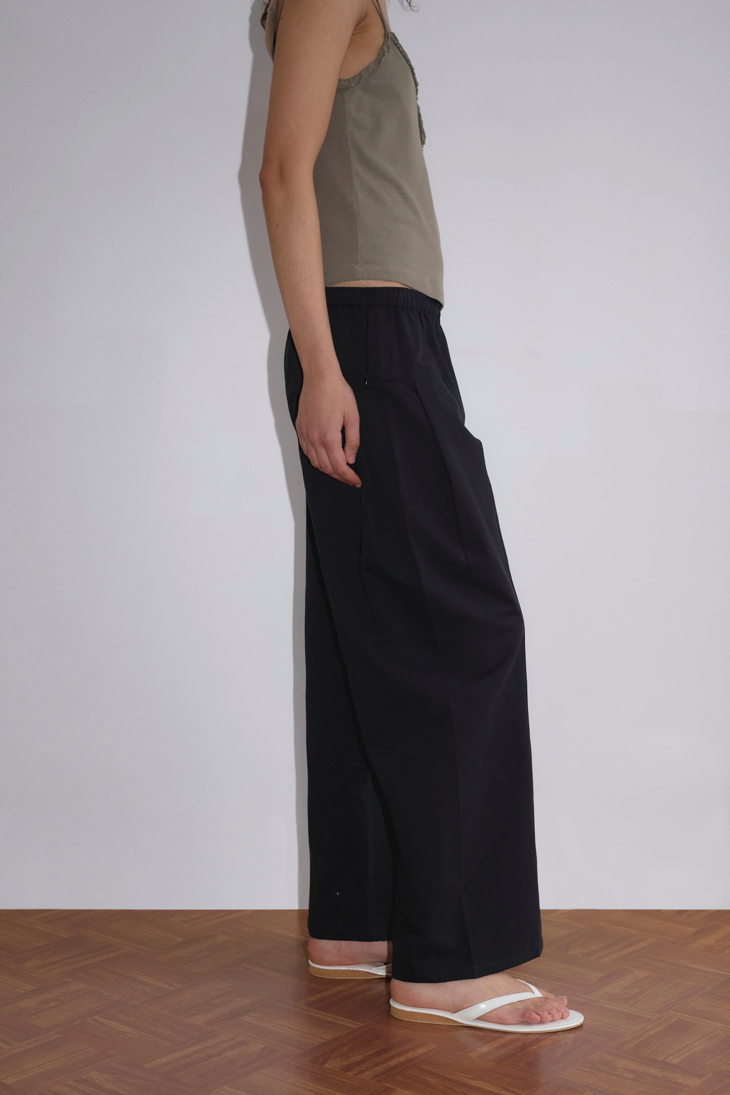 A person stands on a wooden floor against a plain white wall, wearing a sleeveless beige top, Pleat Pant - Black, and white flip-flops. Only the side of their body is visible from head to feet.