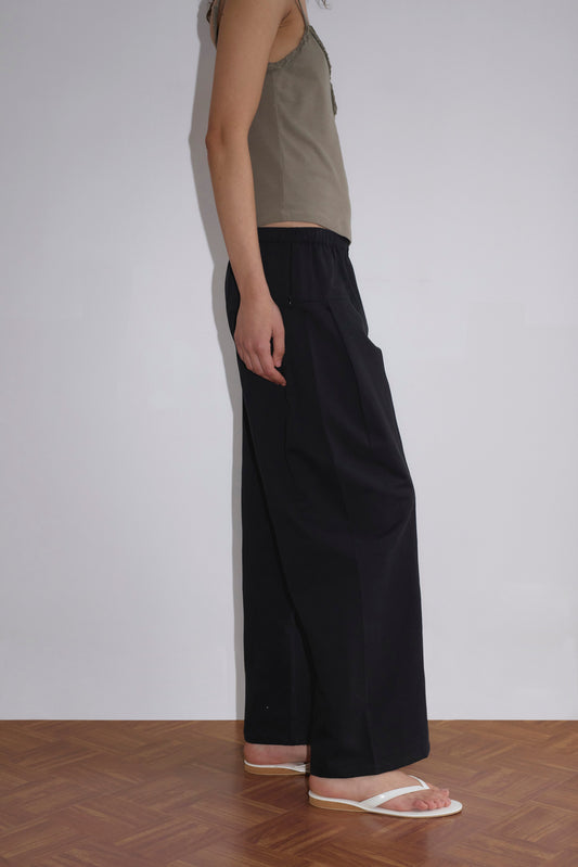 A person stands on a wooden floor against a plain white wall, wearing a sleeveless beige top, Pleat Pant - Black, and white flip-flops. Only the side of their body is visible from head to feet.