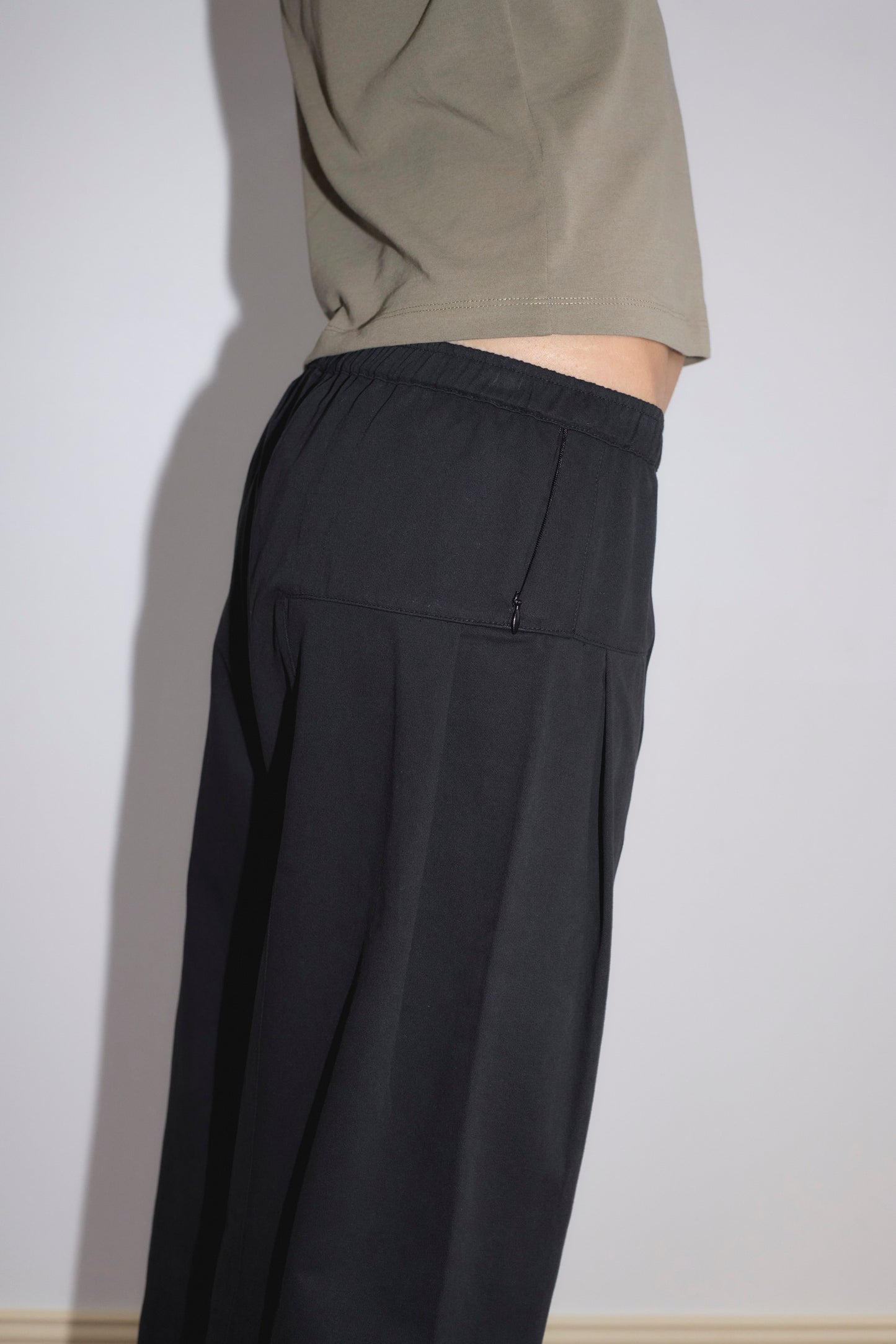 A person stands sideways against a light wall, wearing the Pleat Pant - Black from Deiji Studios. The photo shows only the torso and upper legs, emphasizing the pant's loose fit and elastic waistband.