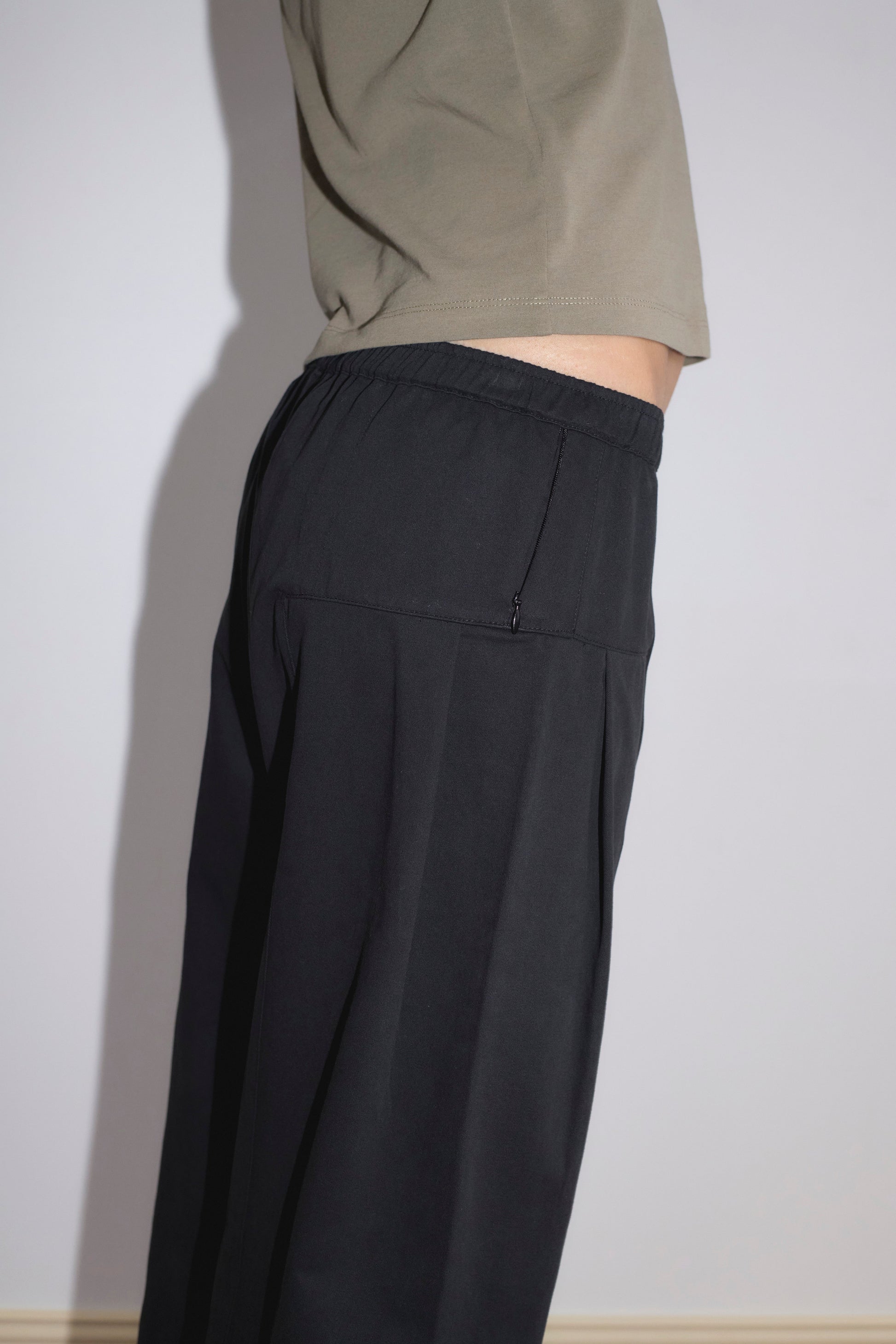 A person stands sideways against a light wall, wearing the Pleat Pant - Black from Deiji Studios. The photo shows only the torso and upper legs, emphasizing the pant's loose fit and elastic waistband.