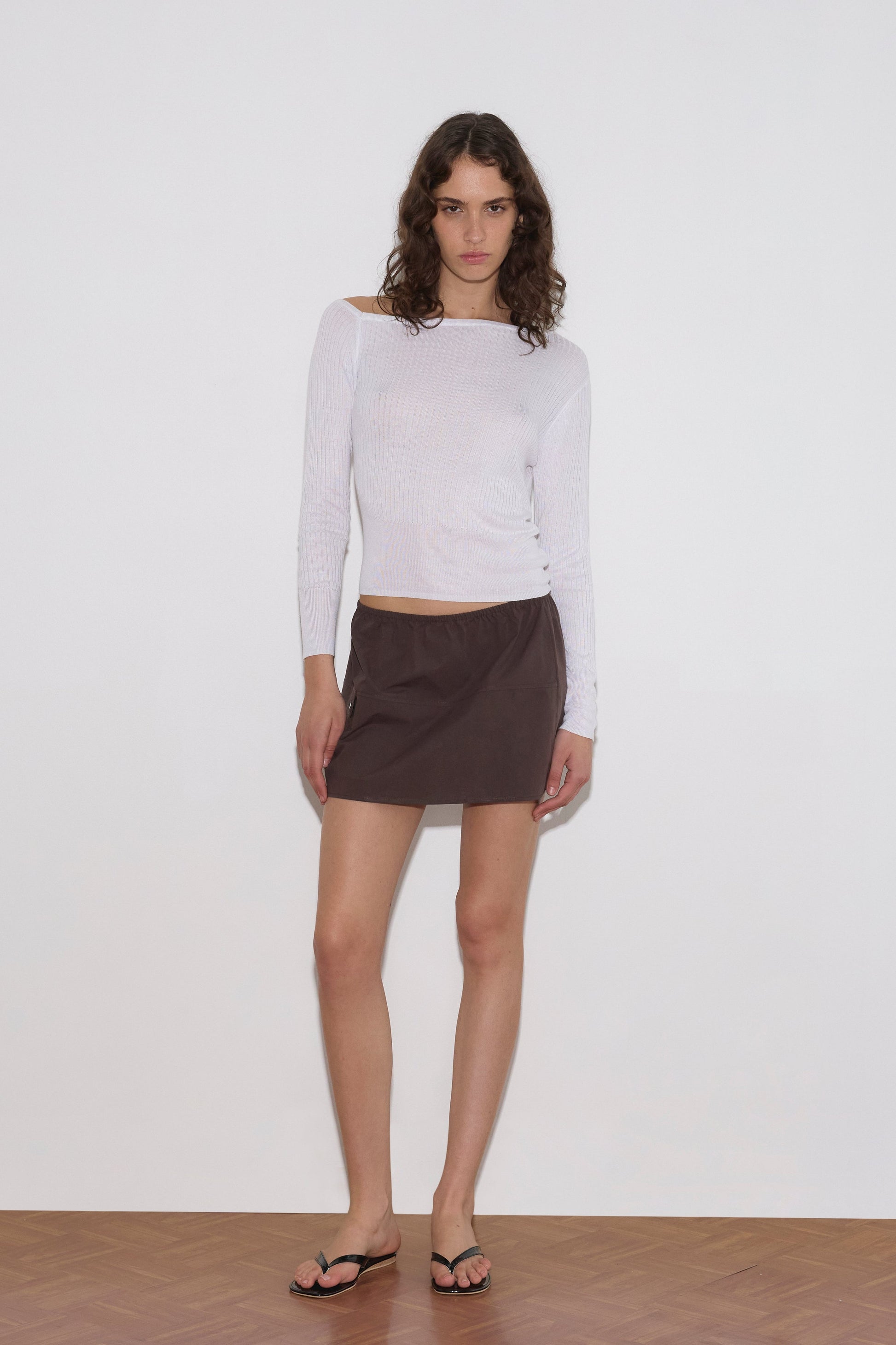 A person with long curly hair stands against a plain white wall on a wooden floor, wearing a white long-sleeve top, black flip-flops, and the Deiji Studios Pocket Skirt - Sepia in mini length.
