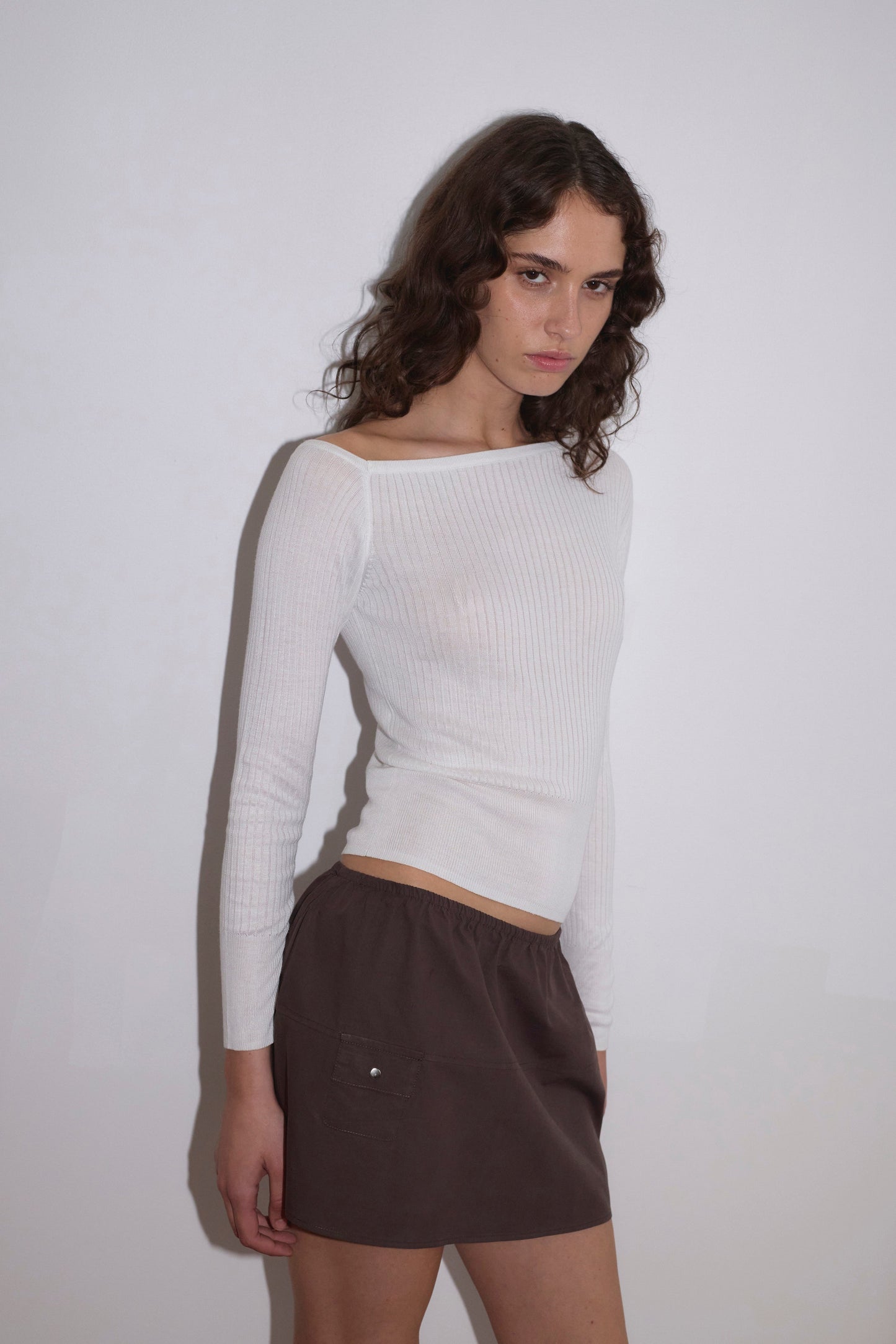A woman with wavy brown hair stands against a plain white wall, wearing the Shoulder Knit Top in white and a brown mini skirt with a side pocket, looking at the camera with a neutral expression.