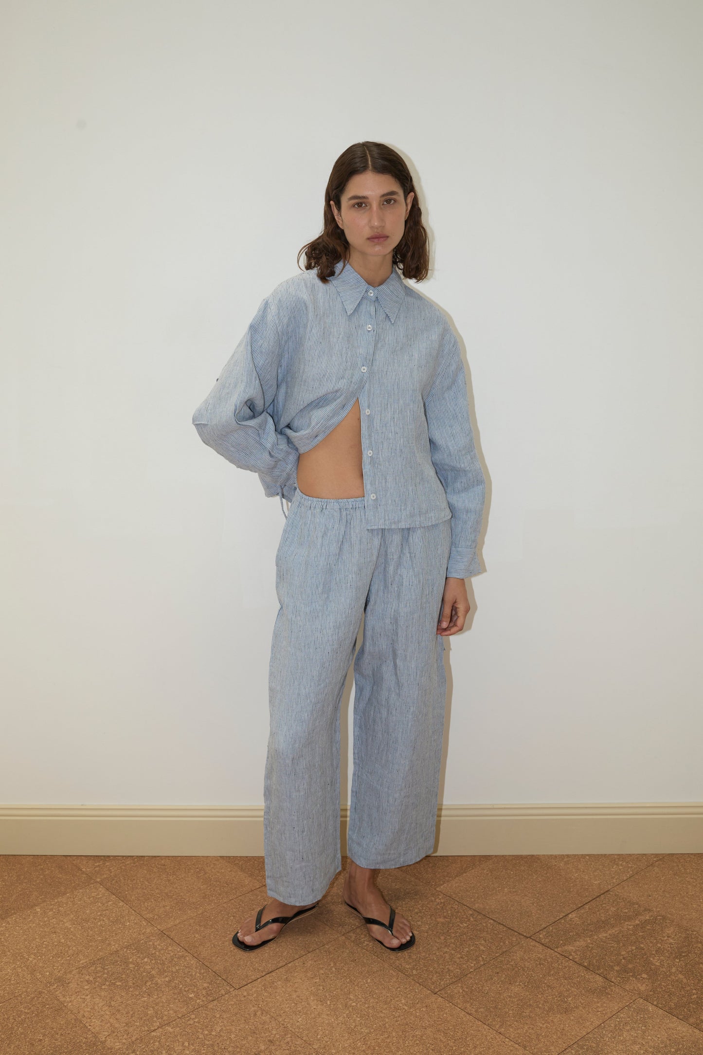 A person with shoulder-length brown hair stands against a white wall, wearing Deiji Studios' 4 Tie Set - Blue Stripe: a loose, patterned French linen matching shirt and pants set, with one side of the organic shirt tucked up, paired with black flip-flops.