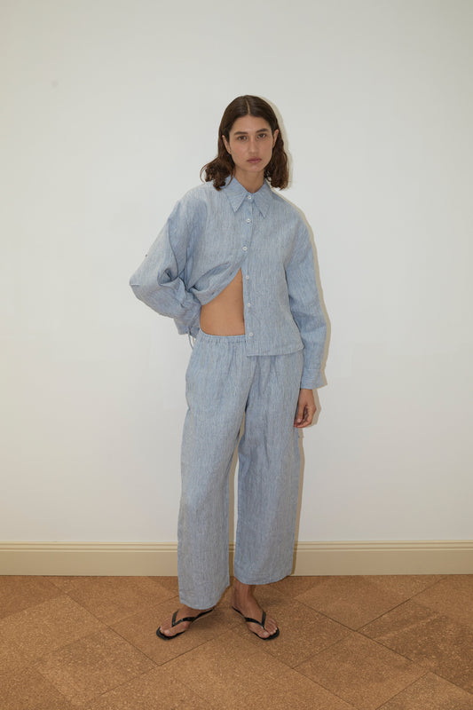 A person with shoulder-length brown hair stands against a white wall, wearing Deiji Studios' 4 Tie Set - Blue Stripe: a loose, patterned French linen matching shirt and pants set, with one side of the organic shirt tucked up, paired with black flip-flops.