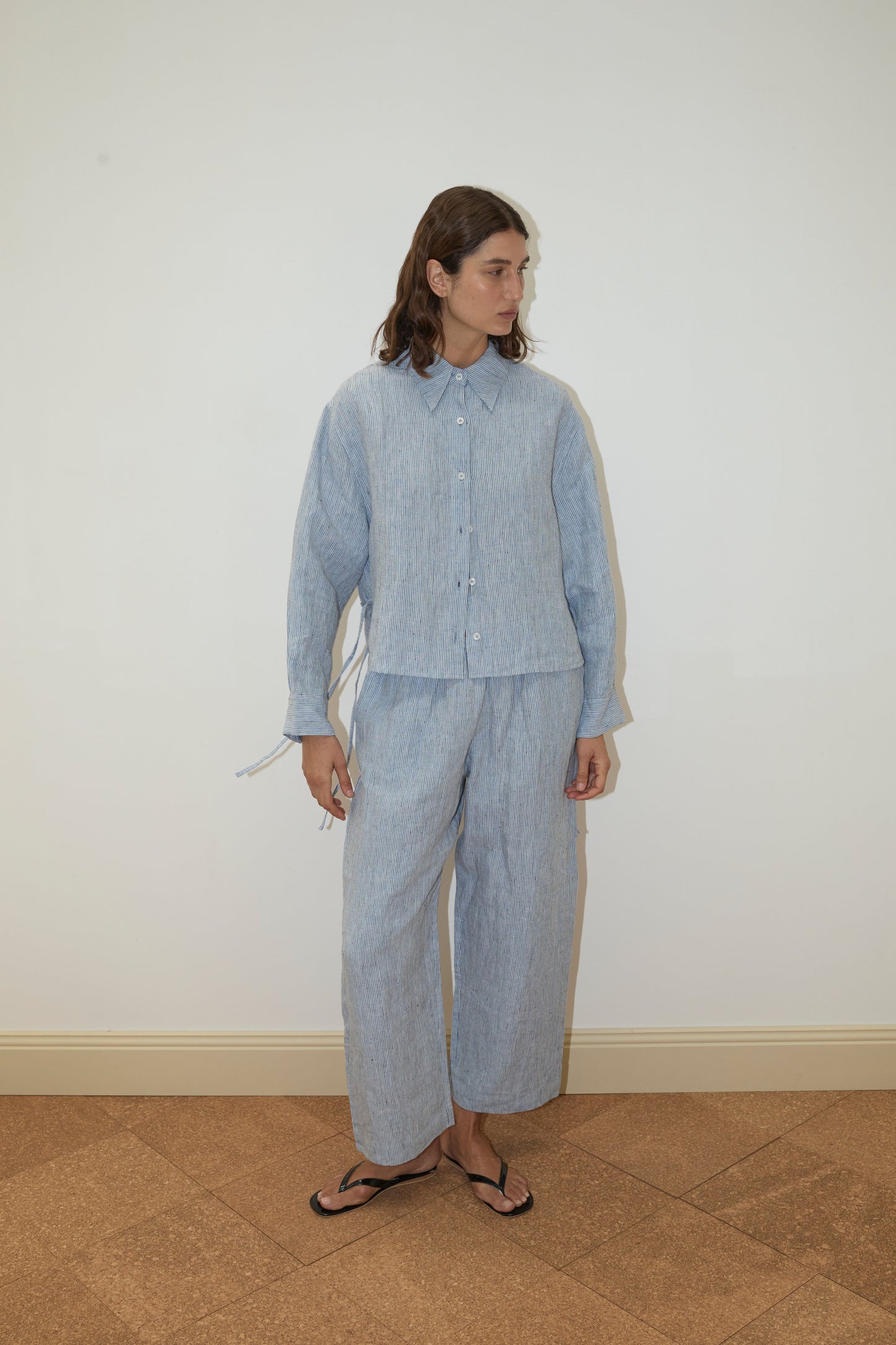 A person with long brown hair stands indoors against a plain white wall, wearing a 4 Tie Set - Blue Stripe.
