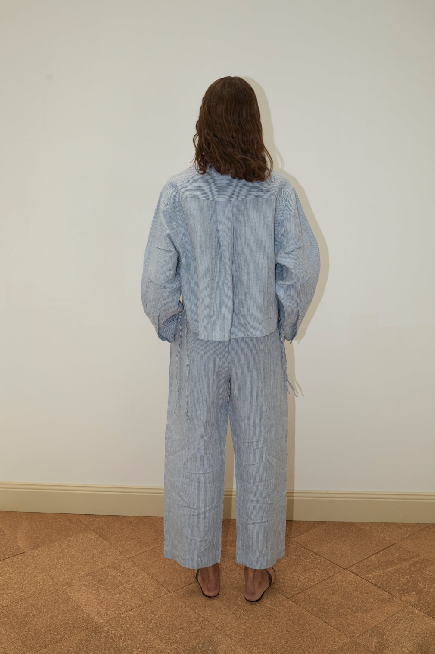 A person with wavy brown hair faces a white wall, wearing Deiji Studios' 4 Tie Set - Blue Stripe: a loose, light blue French linen top and wide-leg pants, paired with open-toe sandals. Their back is to the camera.