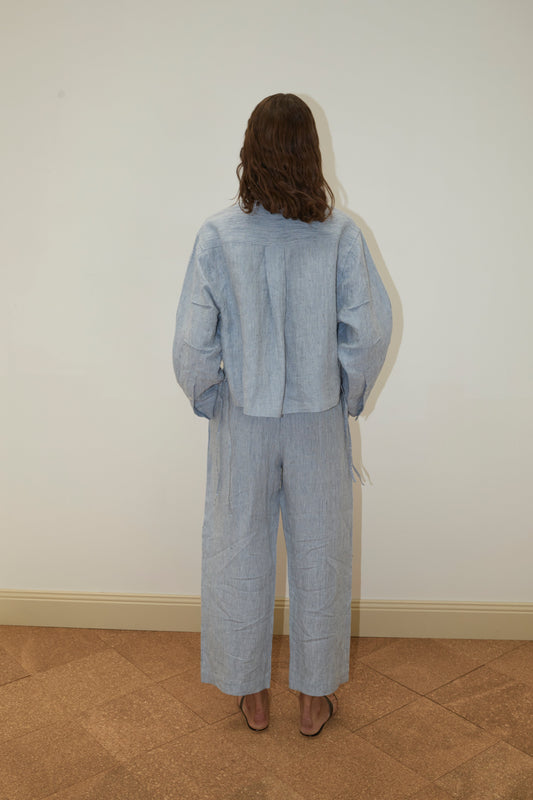 A person with wavy brown hair faces a white wall, wearing Deiji Studios' 4 Tie Set - Blue Stripe: a loose, light blue French linen top and wide-leg pants, paired with open-toe sandals. Their back is to the camera.