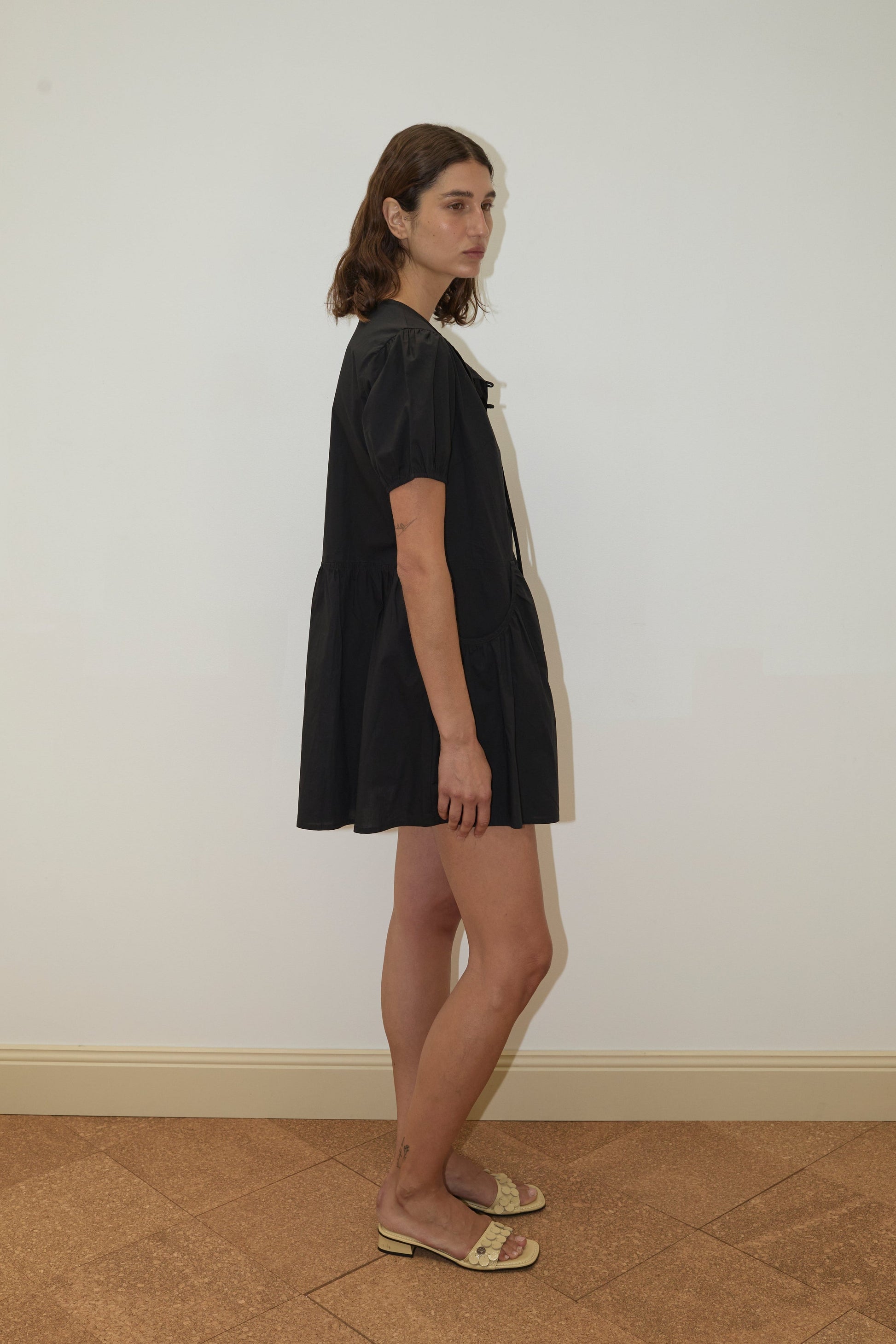 A woman with shoulder-length brown hair stands sideways in the Pocket Mini Dress - Black from Deiji Studios and beige open-toe sandals, set against a plain white wall and tan tiled floor.