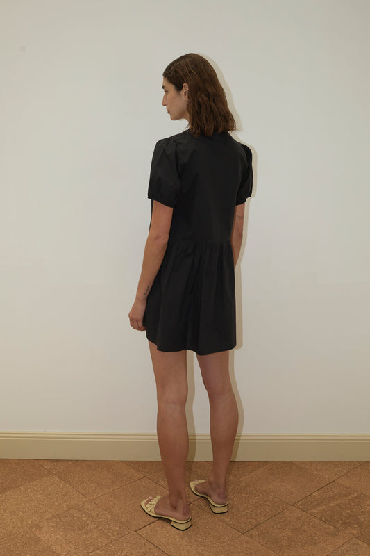 A person with shoulder-length brown hair wears the Pocket Mini Dress - Black from Deiji Studios and beige sandals. They stand indoors on a tiled floor near a cream-colored wall with a baseboard, facing away from the camera.