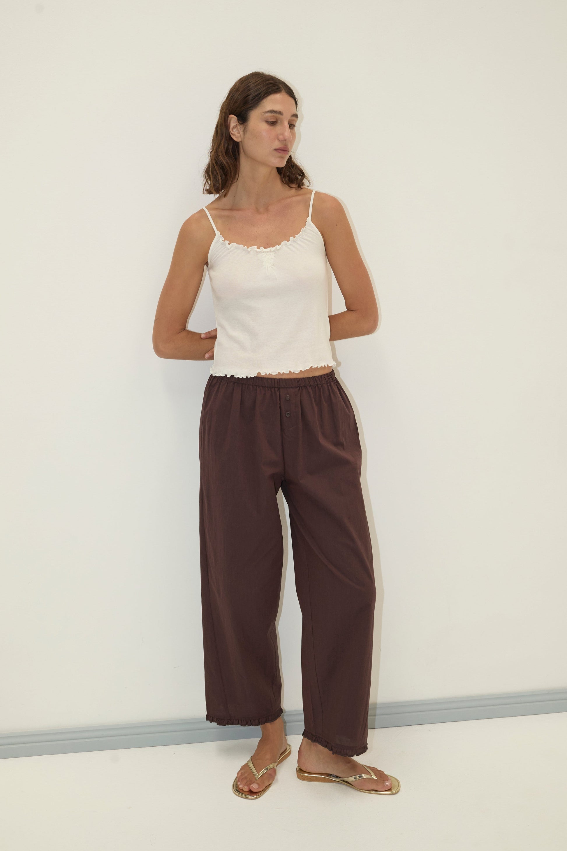 A woman with long wavy hair stands against a white wall, wearing the Flounce Tank - Cream by Deiji Studios, loose dark brown pants, and clear sandals. She looks slightly to the side with her hands in her pockets.